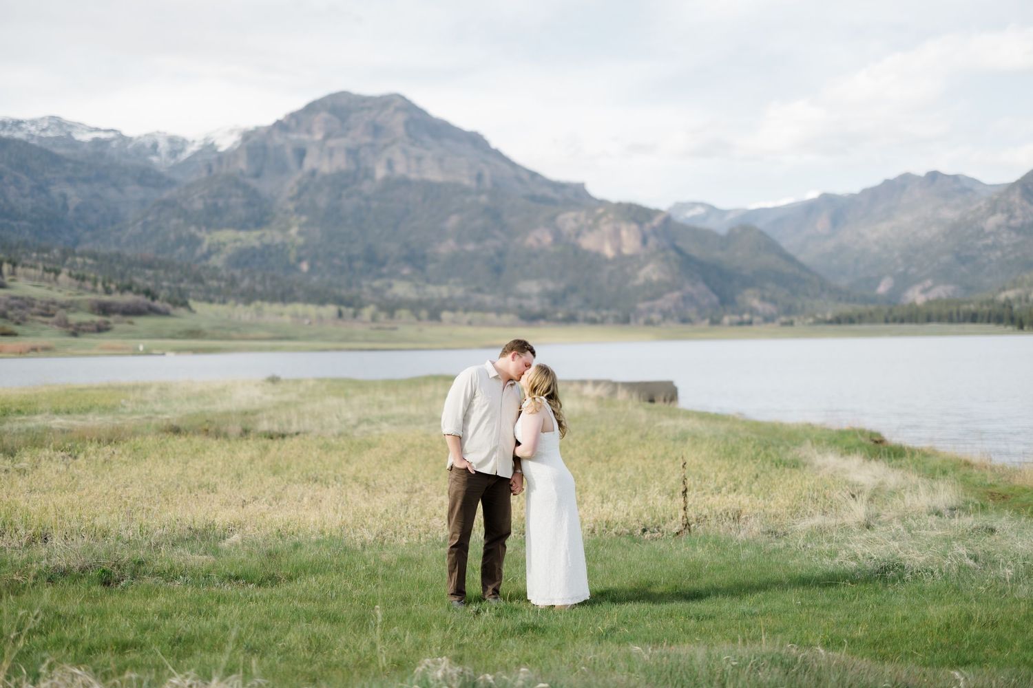 Couple embracing and kissing in a grassy field with a scenic mountain lake behind them.