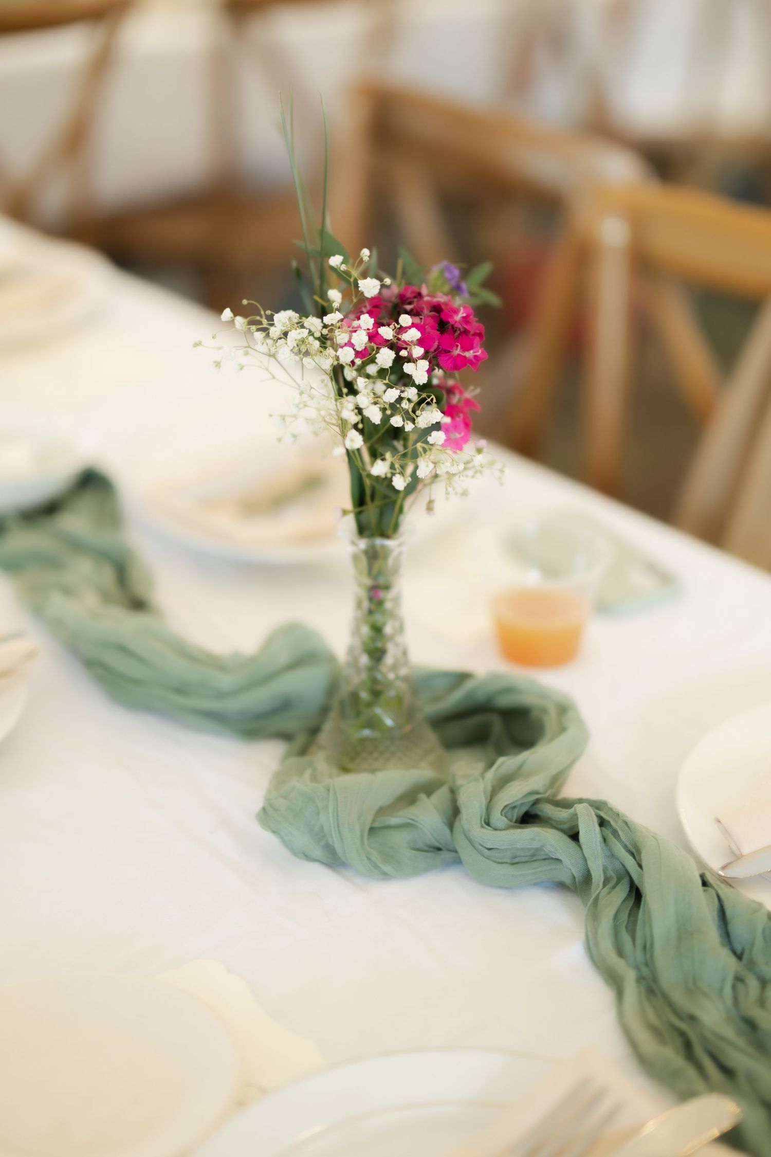 Elegant wedding table centerpiece with pink flowers and baby's breath in glass vase on sage green cheesecloth runner.