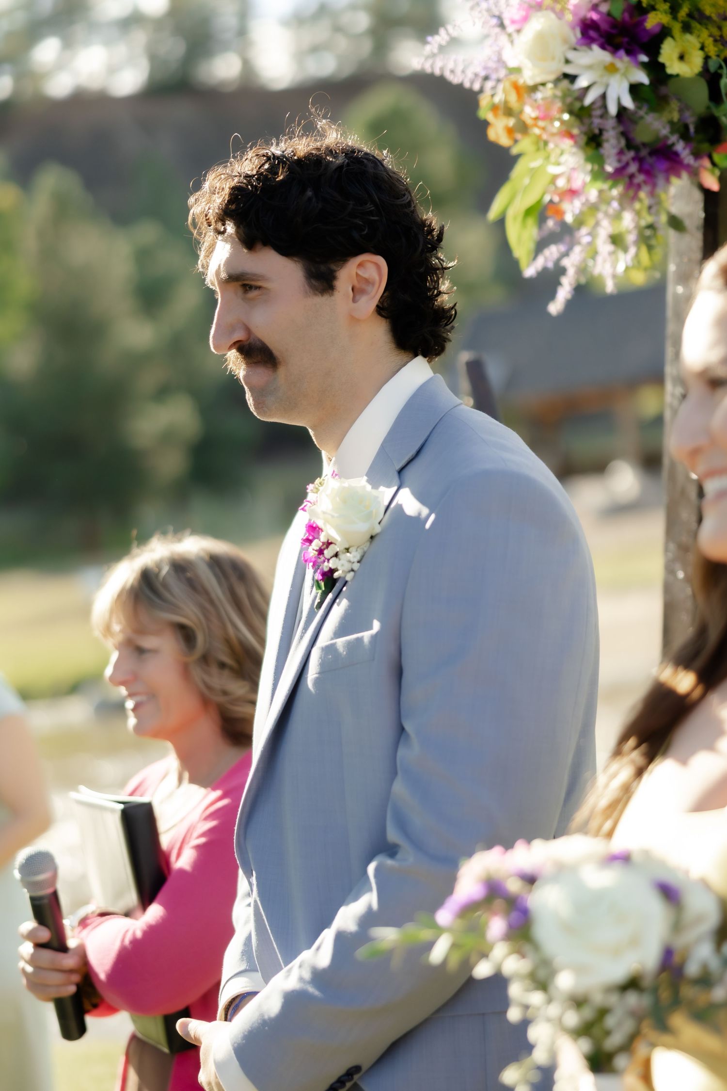 Groom in gray suit with white boutonniere smiling at outdoor wedding ceremony with flowers and officiant.