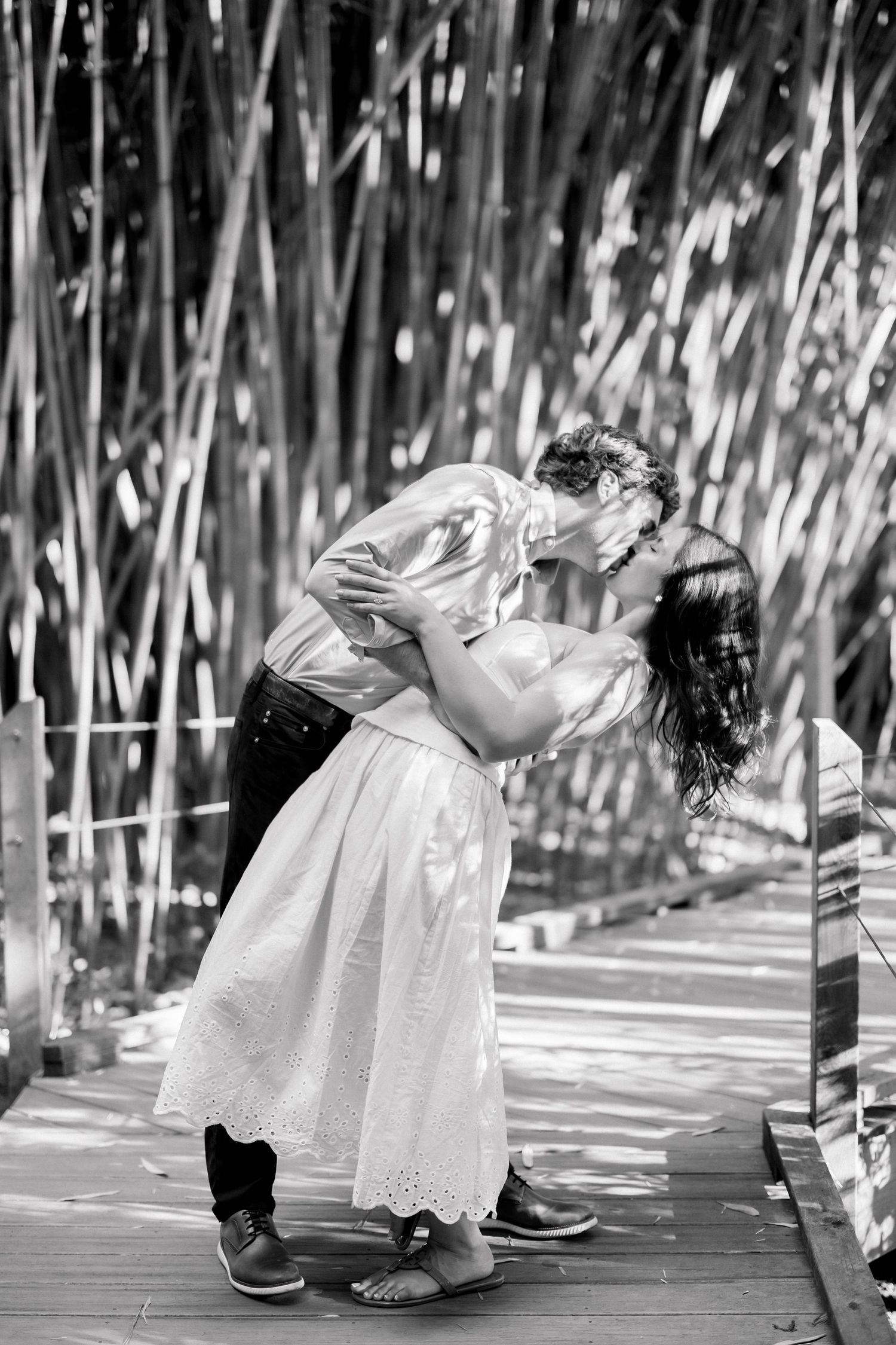Black and white photo of a couple sharing a romantic kiss on a wooden walkway surrounded by tall bamboo.