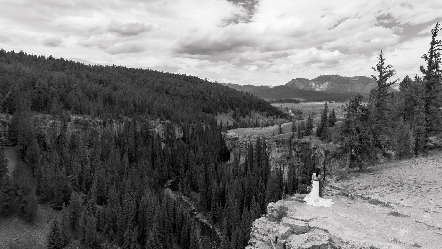 Black and white aerial photo of a wedding couple standing on a rocky overlook surrounded by dense pine forests and mountains.