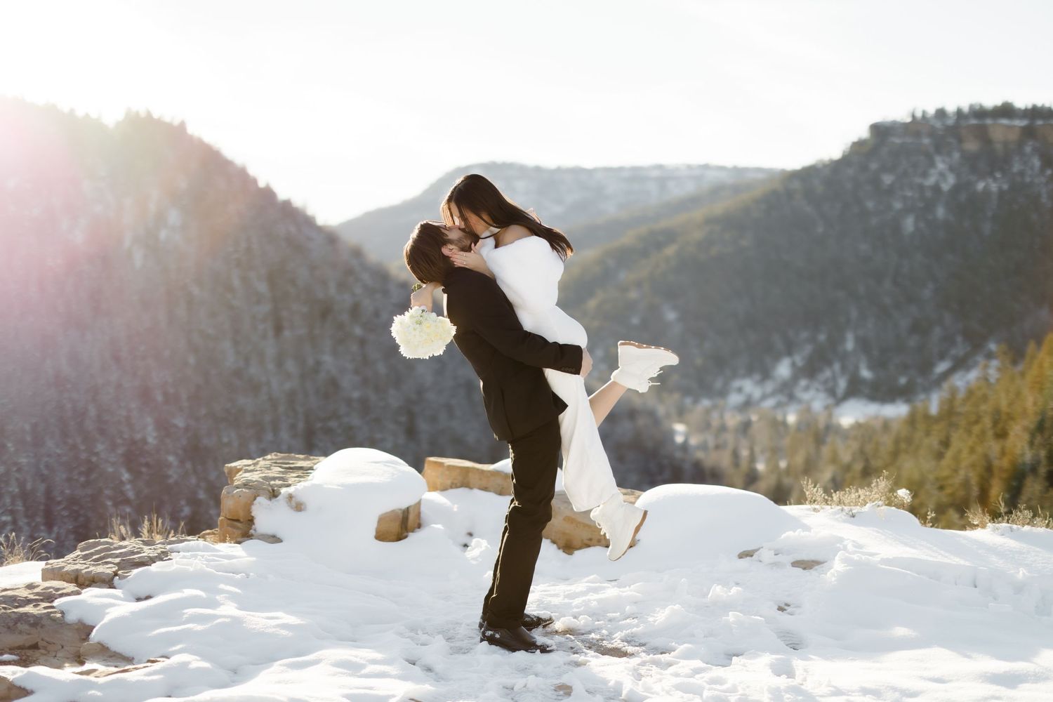 Groom lifting bride in snowy mountain landscape during winter wedding photoshoot with sunlit peaks behind.