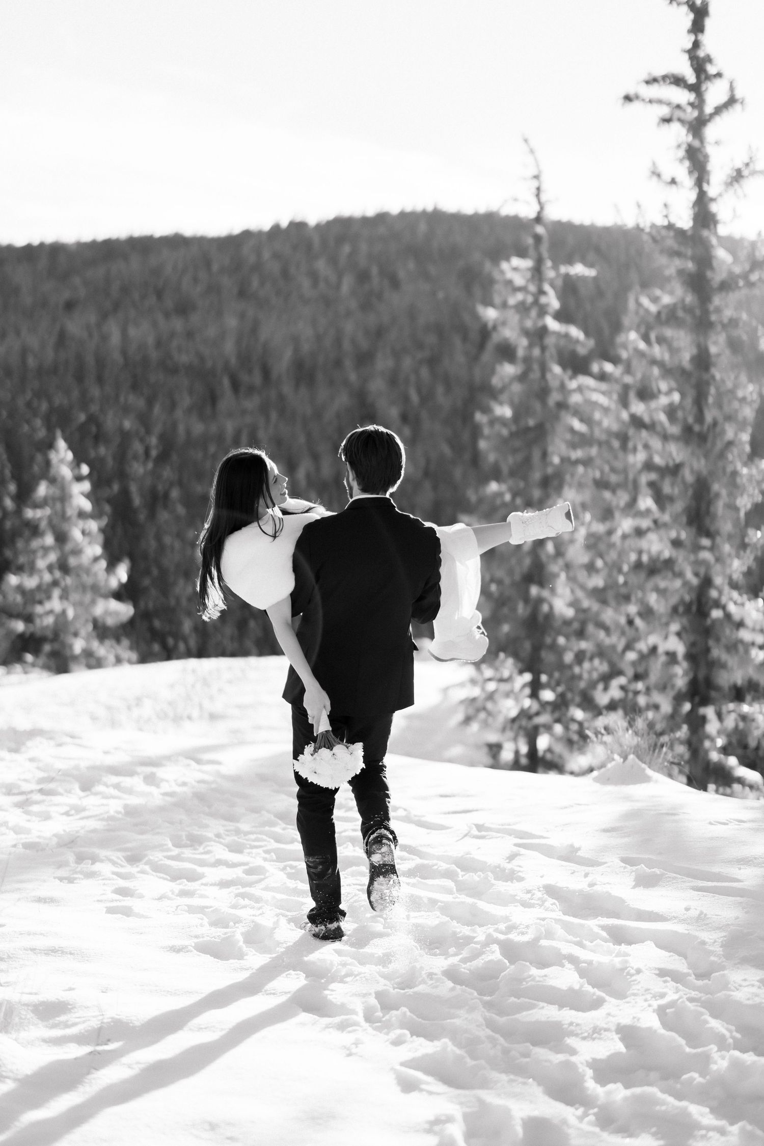 Black and white photo of groom carrying bride through snowy mountain forest.