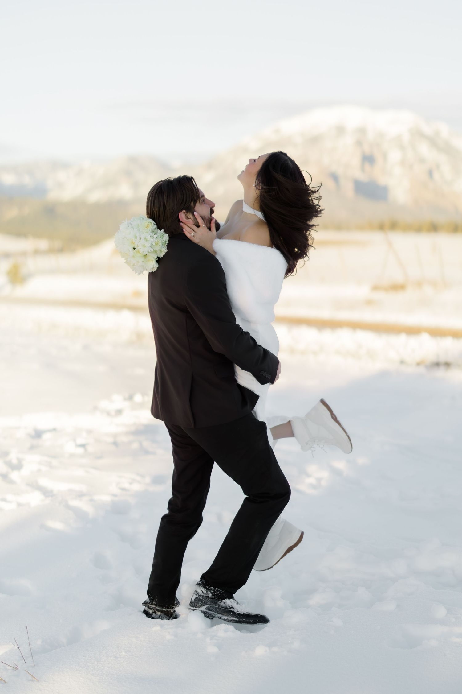 Groom lifts joyful bride in white dress as she laughs in snowy winter landscape.