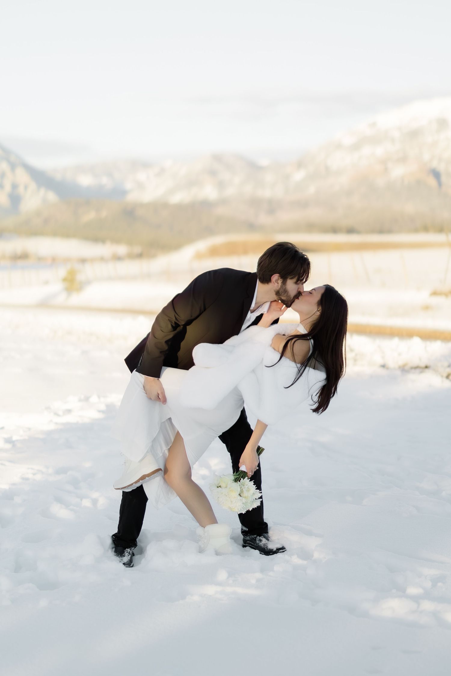 Groom dips bride for a romantic kiss in snowy mountain setting during winter wedding photoshoot.
