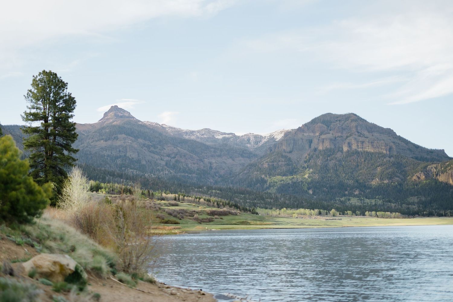 Scenic mountain lake with calm blue waters, pine trees, and rocky peaks under a partly cloudy sky.
