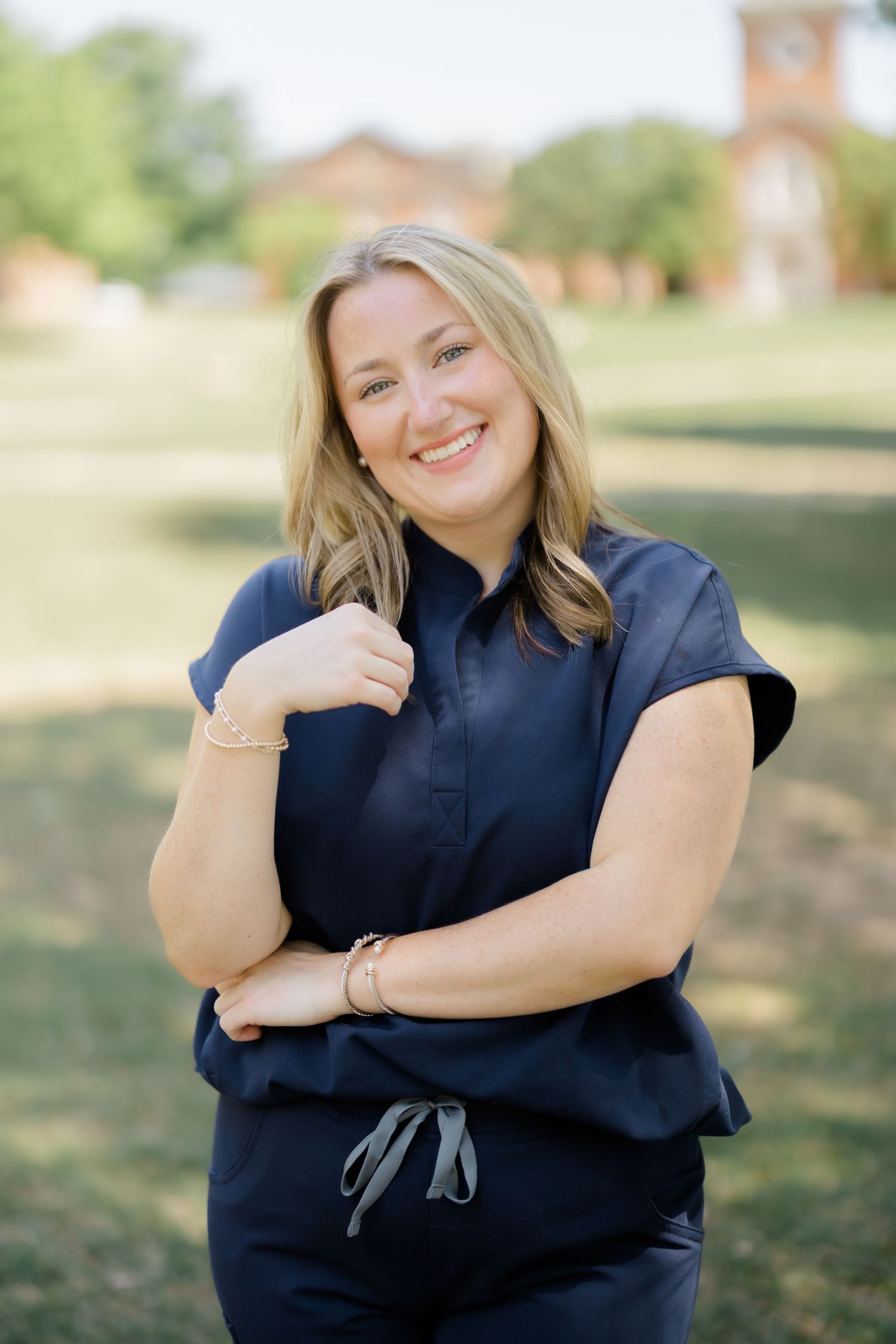 Smiling blonde woman in navy blue scrubs posing outdoors on green lawn with brick building in background.