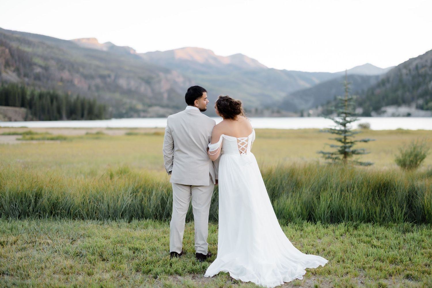 Bride and groom gaze toward mountain lake landscape, bride in flowing white gown with lace-up back detail.