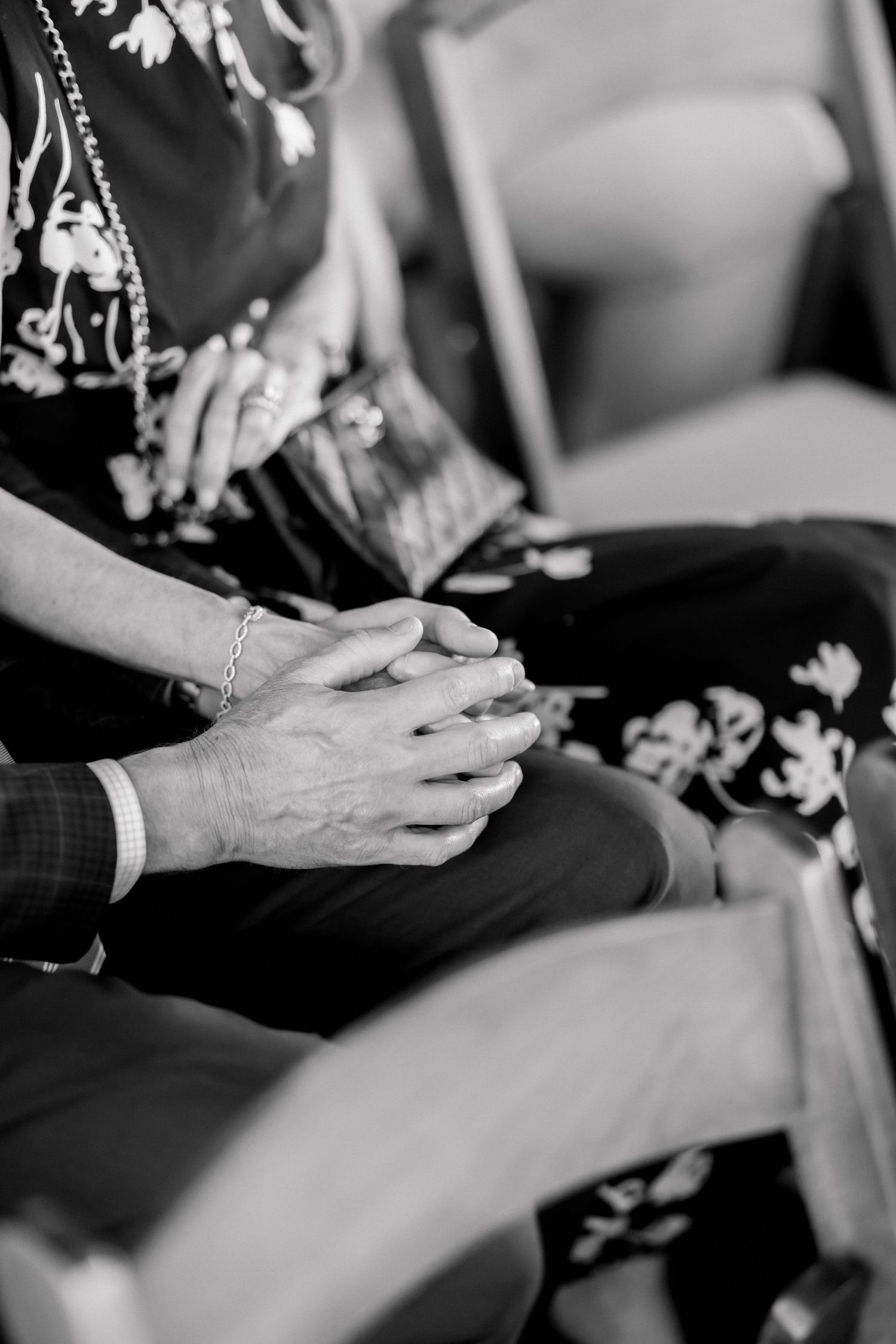 Black and white photo of elderly couple holding hands while seated, wearing elegant attire with floral patterns.