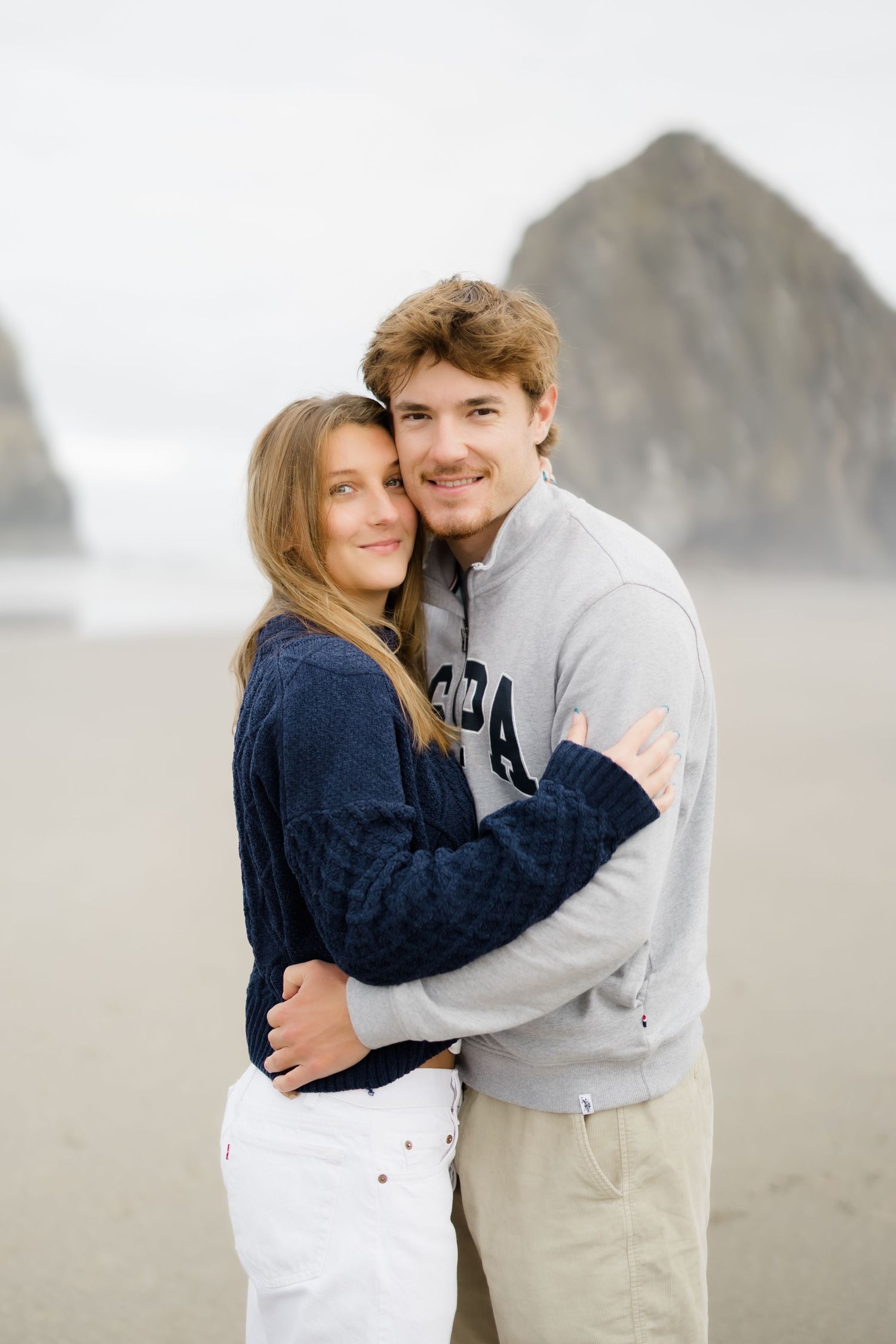 A young couple embraces on a misty beach with a large rock formation in the background, wearing casual sweaters.