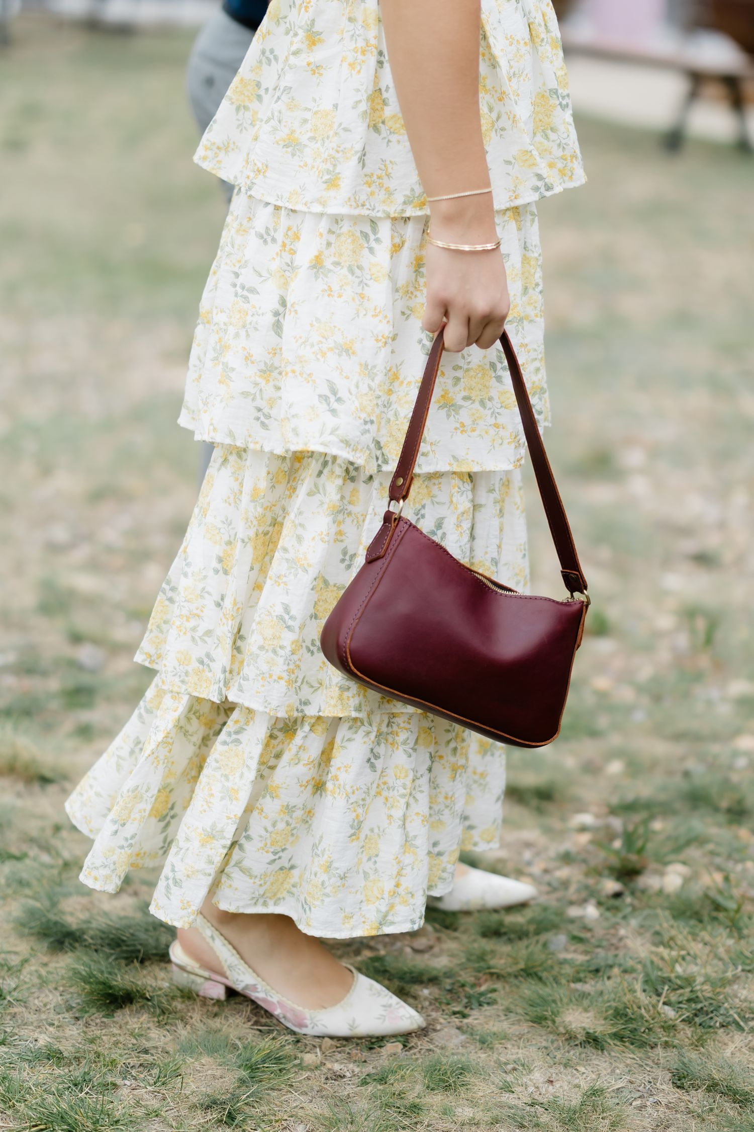Woman in floral tiered maxi dress holding a burgundy leather mini shoulder bag, wearing white kitten heels.
