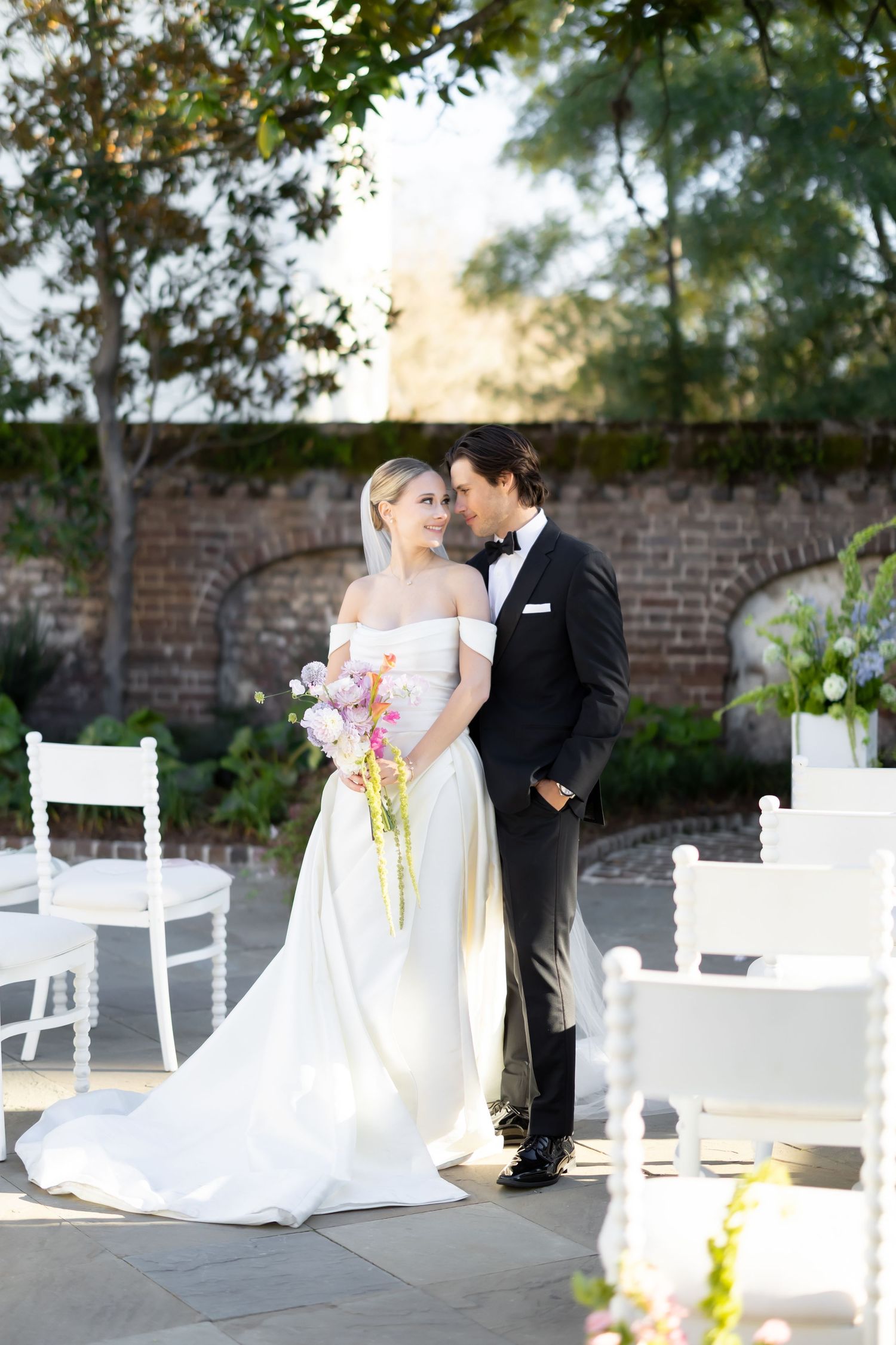 Bride in off-shoulder white gown holds pink bouquet as groom in black tuxedo embraces her at outdoor wedding.