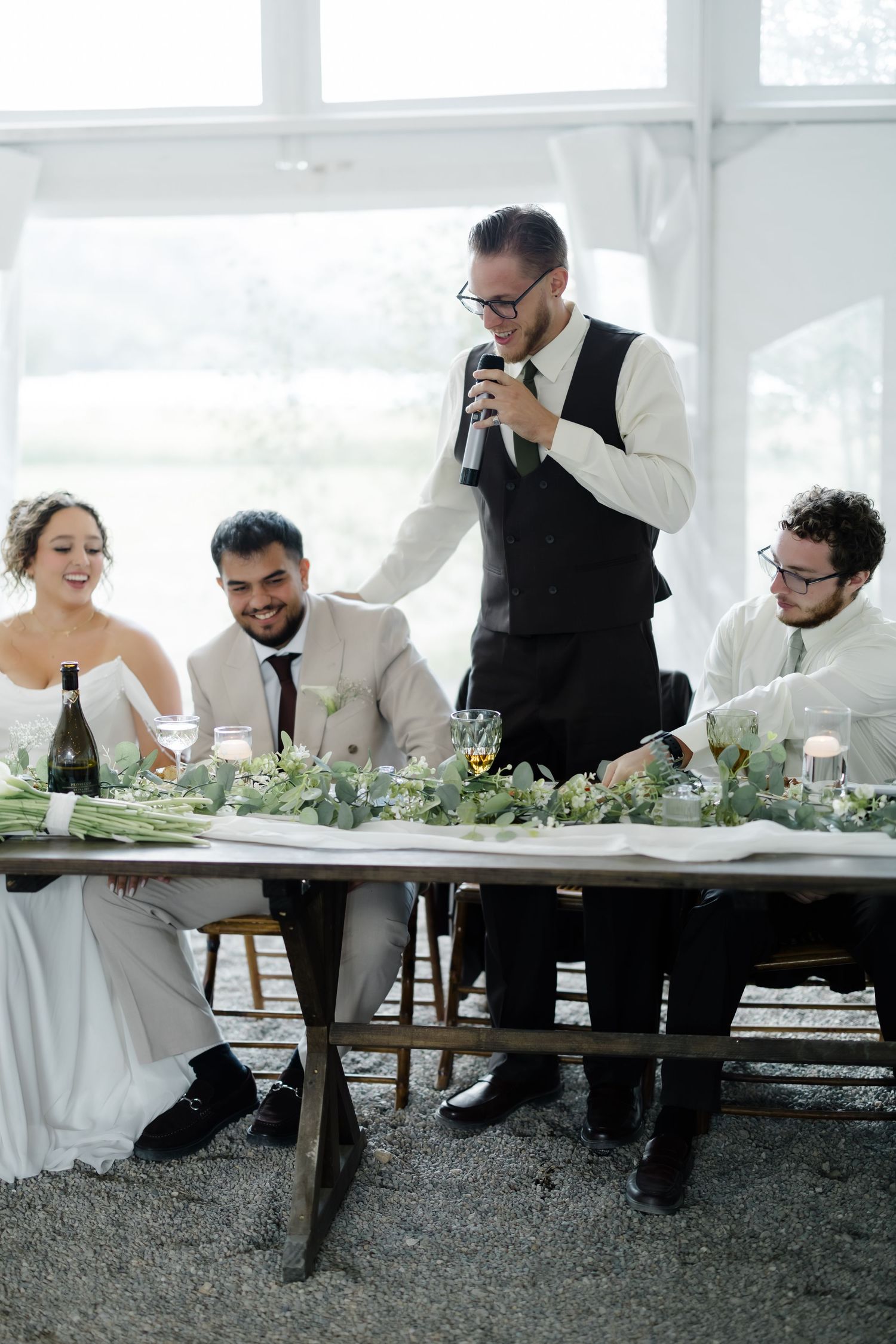 A best man in a vest delivers a heartfelt speech at the wedding head table while guests smile.