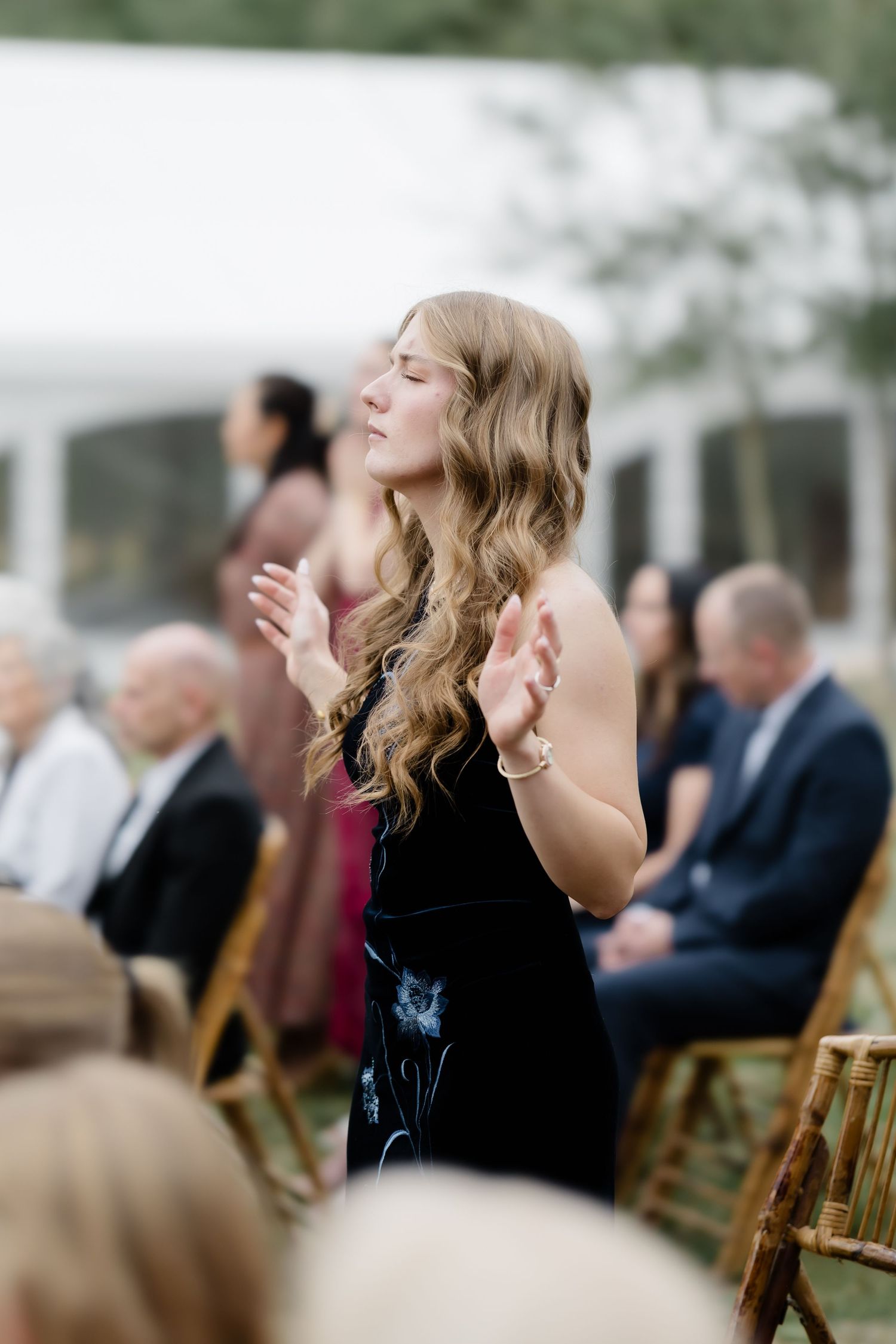 Young woman with wavy hair praying with raised hands during outdoor wedding ceremony.
