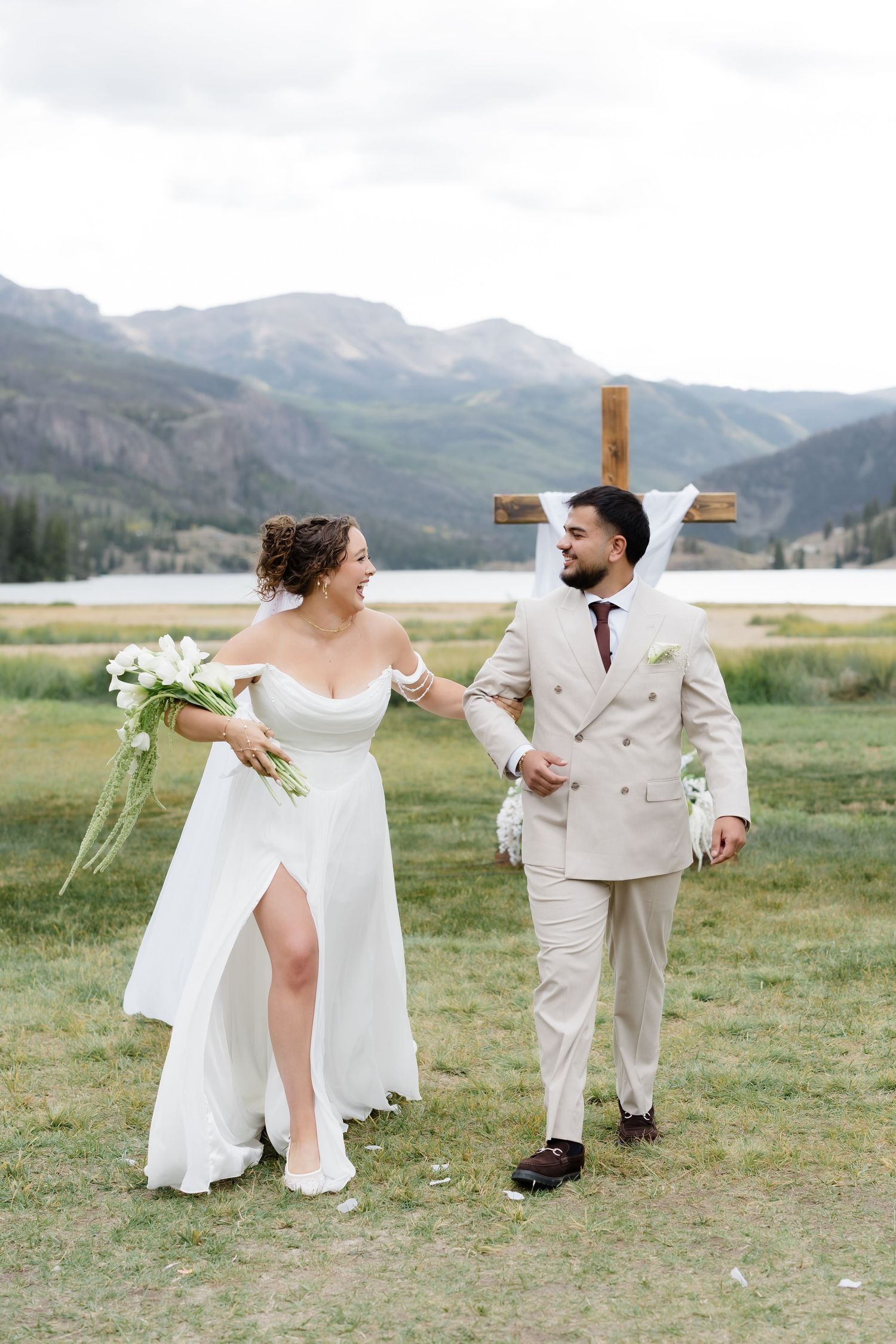 Newlyweds walking together smiling after outdoor mountain wedding ceremony with lake and cross backdrop.