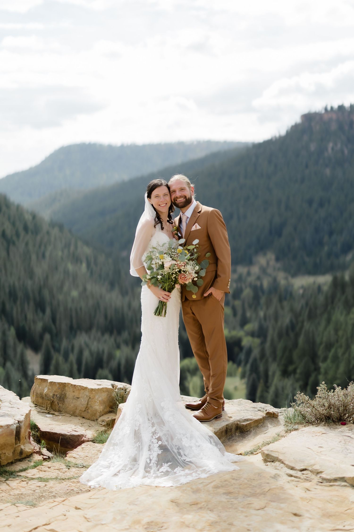 Bride and groom pose on rocky overlook with mountain forest backdrop, she holds wildflower bouquet, he wears brown suit.