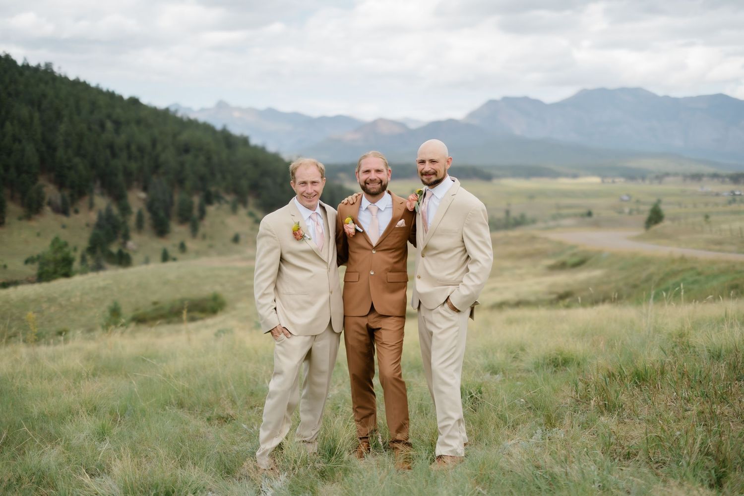 Three men in tan and brown suits pose outdoors at a mountain wedding with scenic Colorado peaks and green meadows behind them.
