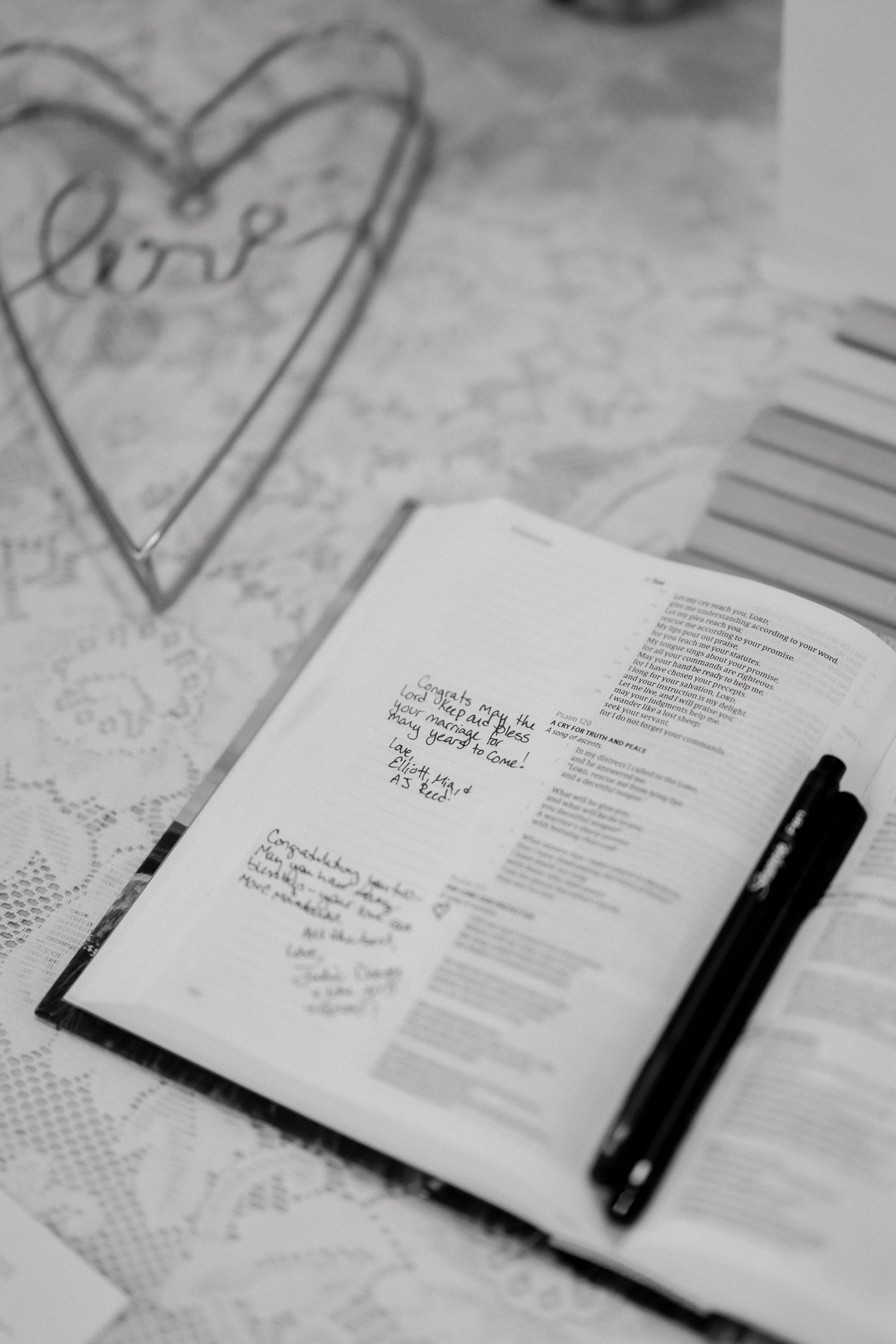 Black and white photo of an open guest book with handwritten notes, pen, and heart-shaped love decoration.