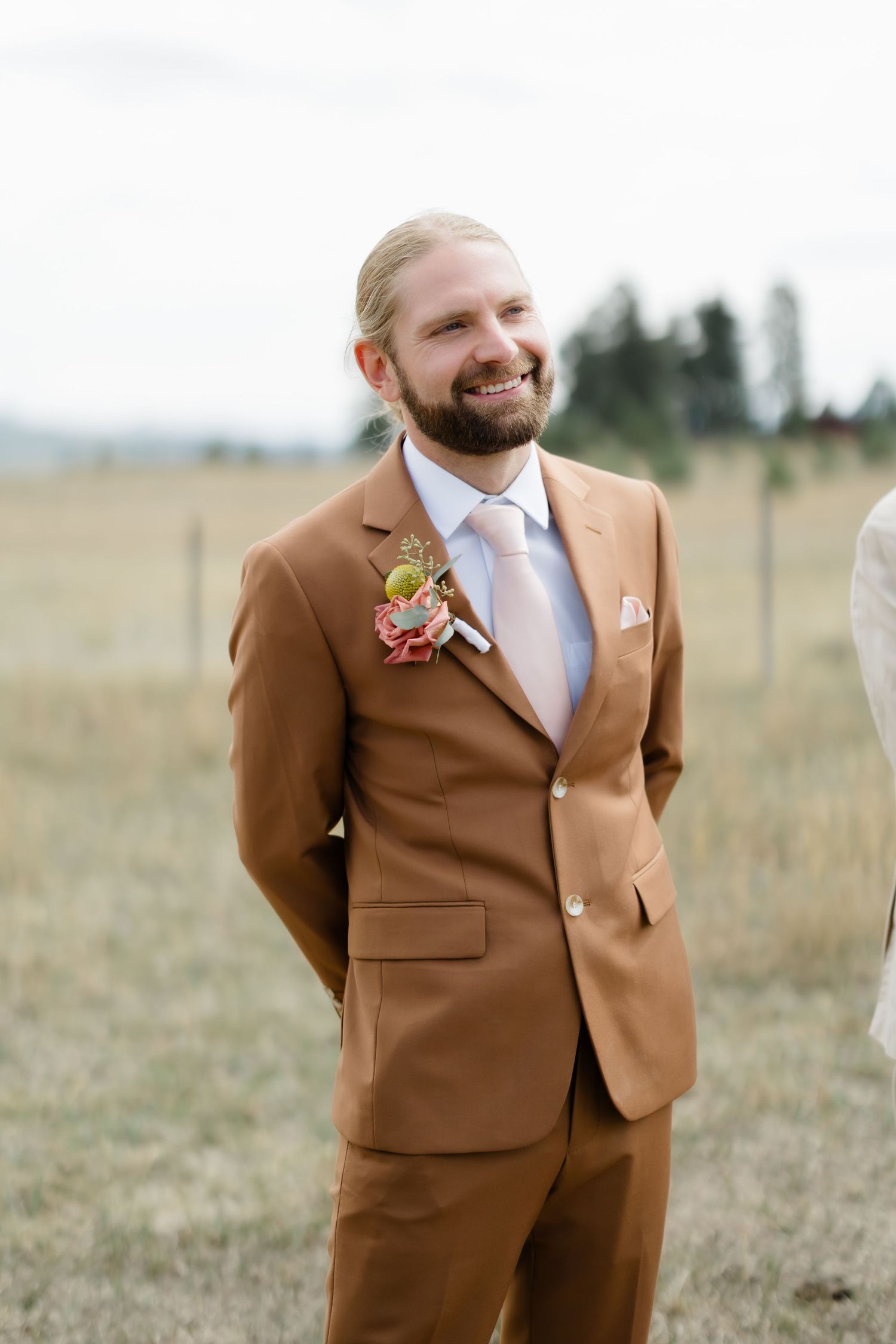 Smiling groom in a tan suit with floral boutonniere at outdoor wedding.