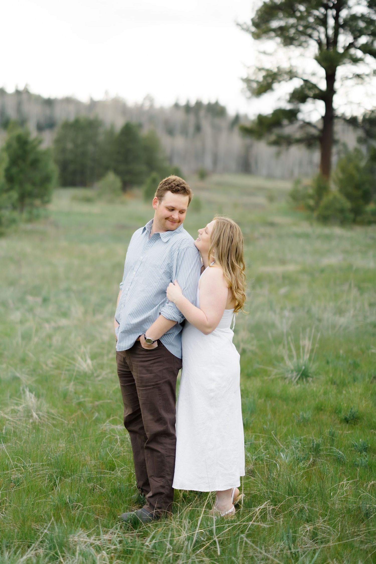 Couple embracing and smiling in a lush green meadow surrounded by pine trees, woman in white dress.