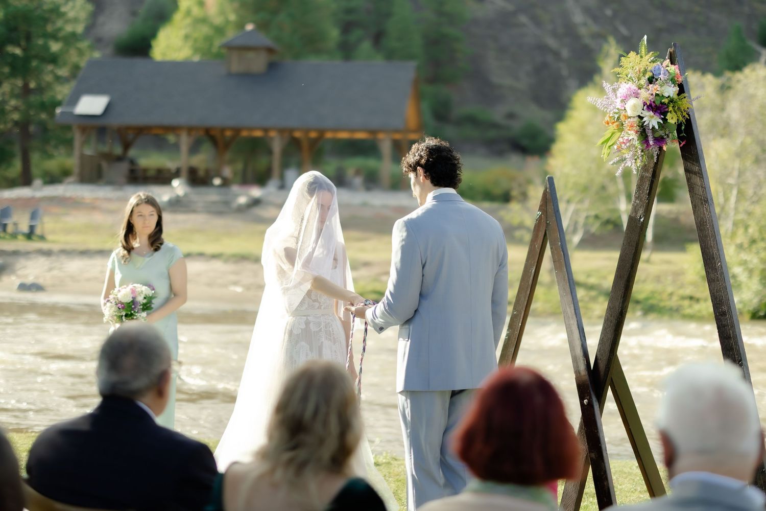 Outdoor wedding ceremony with bride and groom holding hands before a wooden arch adorned with flowers.