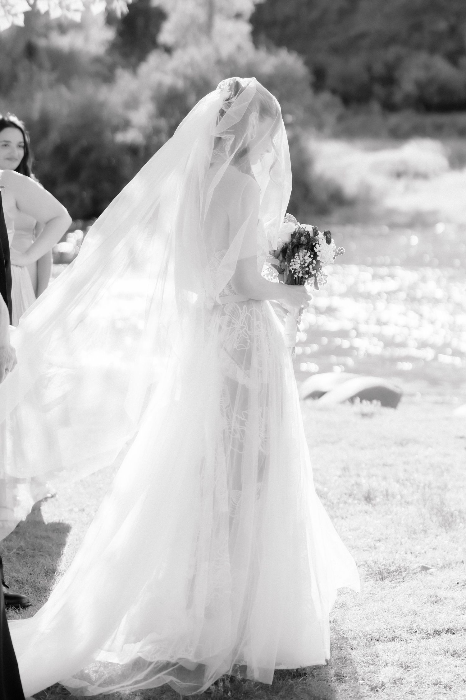 Black and white photo of a bride in a flowing white gown and veil holding a bouquet outdoors near a sunlit river.
