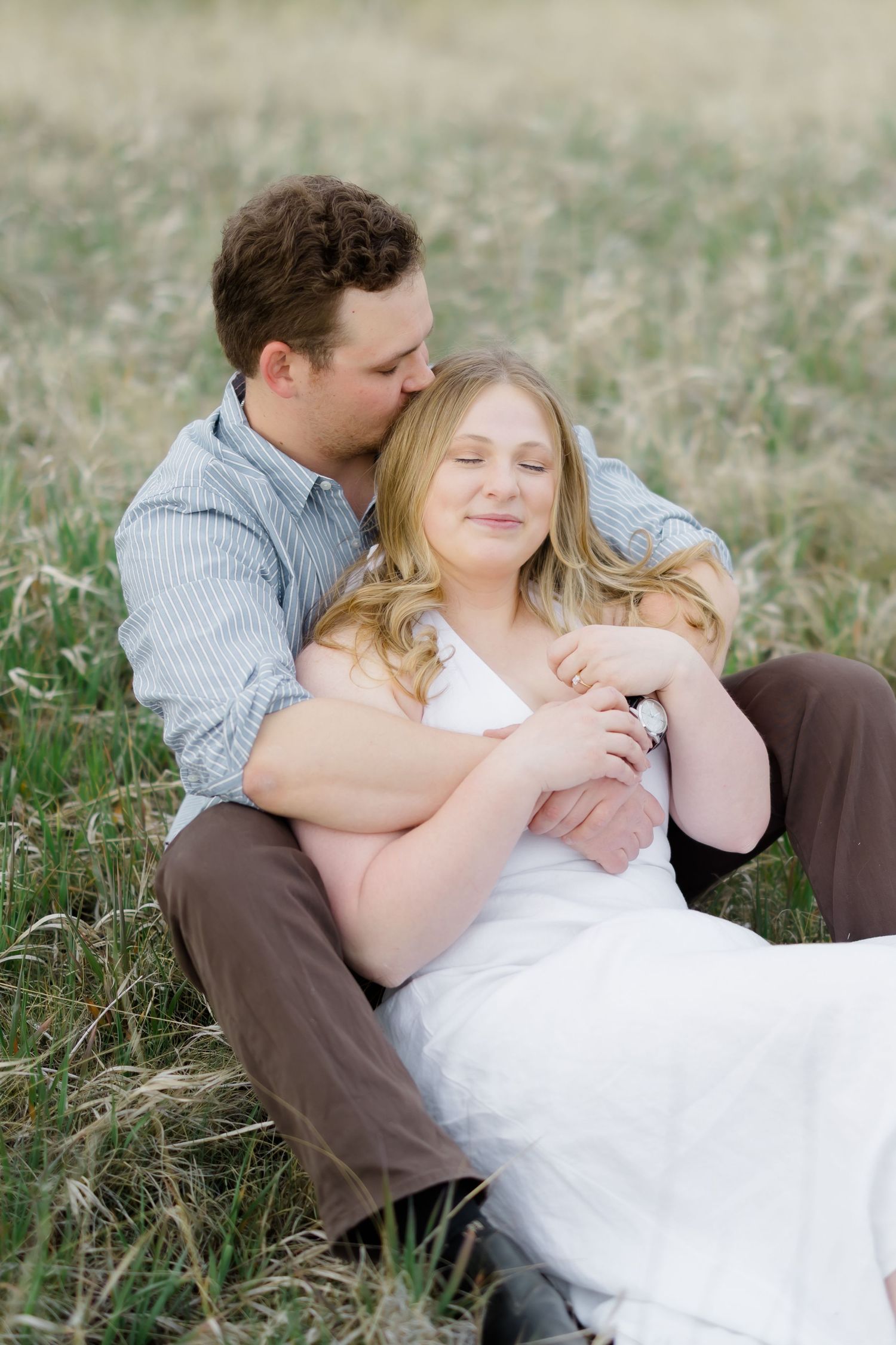 Couple sitting in a grassy field, man embracing woman from behind and kissing her cheek, outdoor engagement photo.