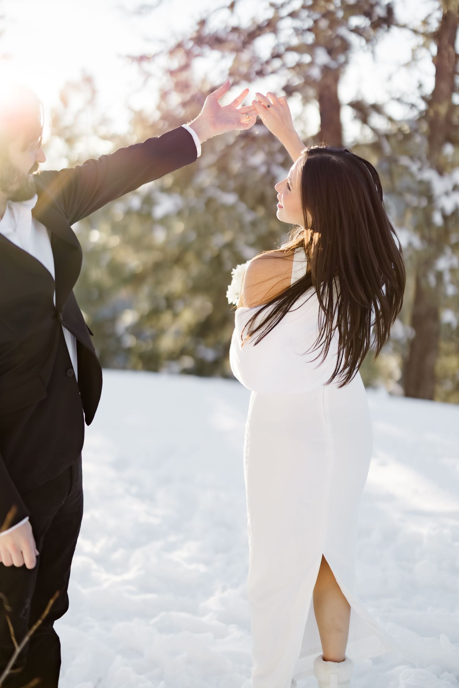 Bride in white gown dancing with groom in black suit in snowy winter wonderland with sunlit trees.