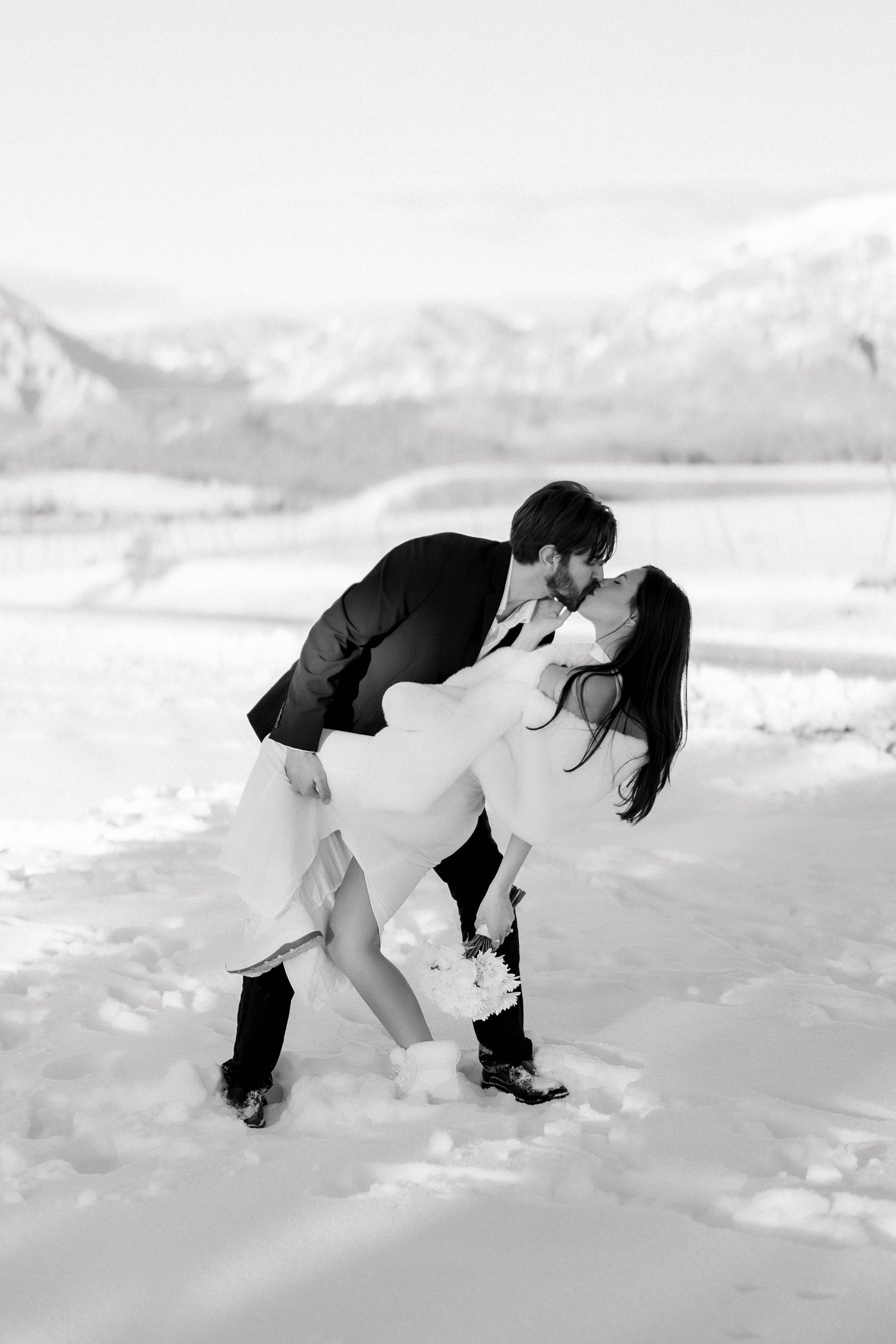 Black and white photo of groom dipping bride for a kiss in a snowy landscape.