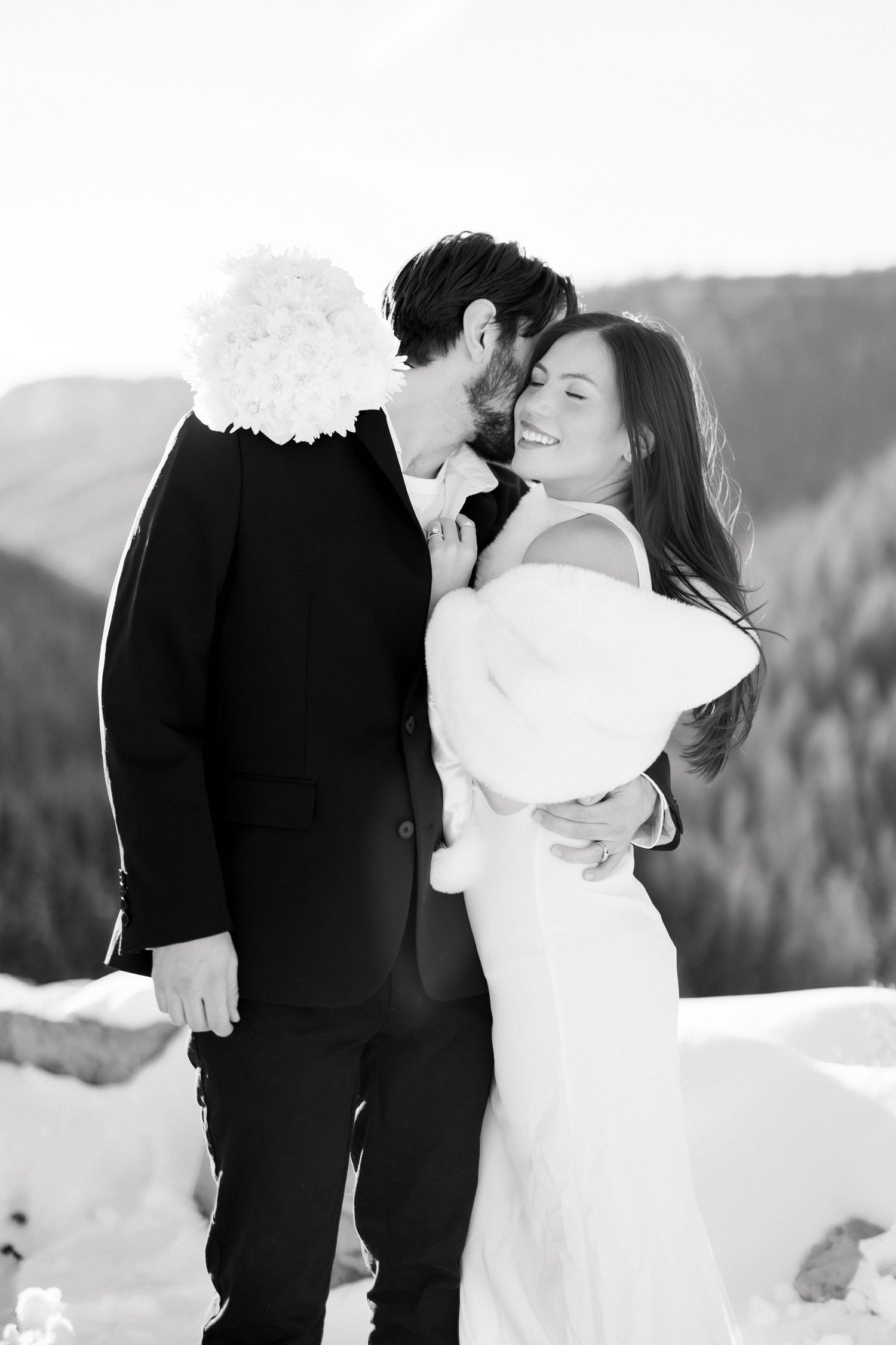 Black and white photo of groom kissing brides cheek as she smiles in snowy mountain landscape.