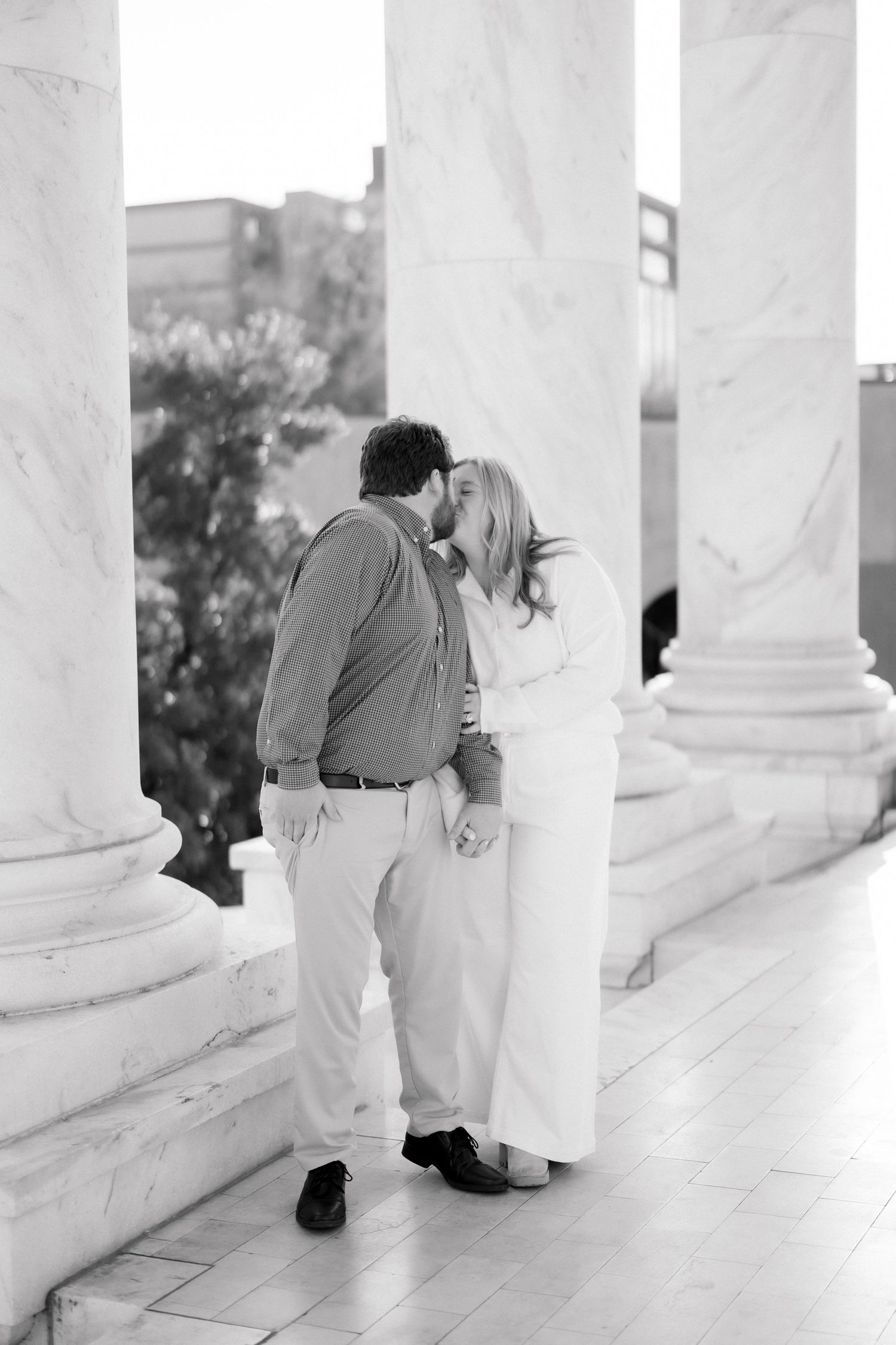 Black and white romantic photo of couple kissing between tall marble columns on a classic building exterior.