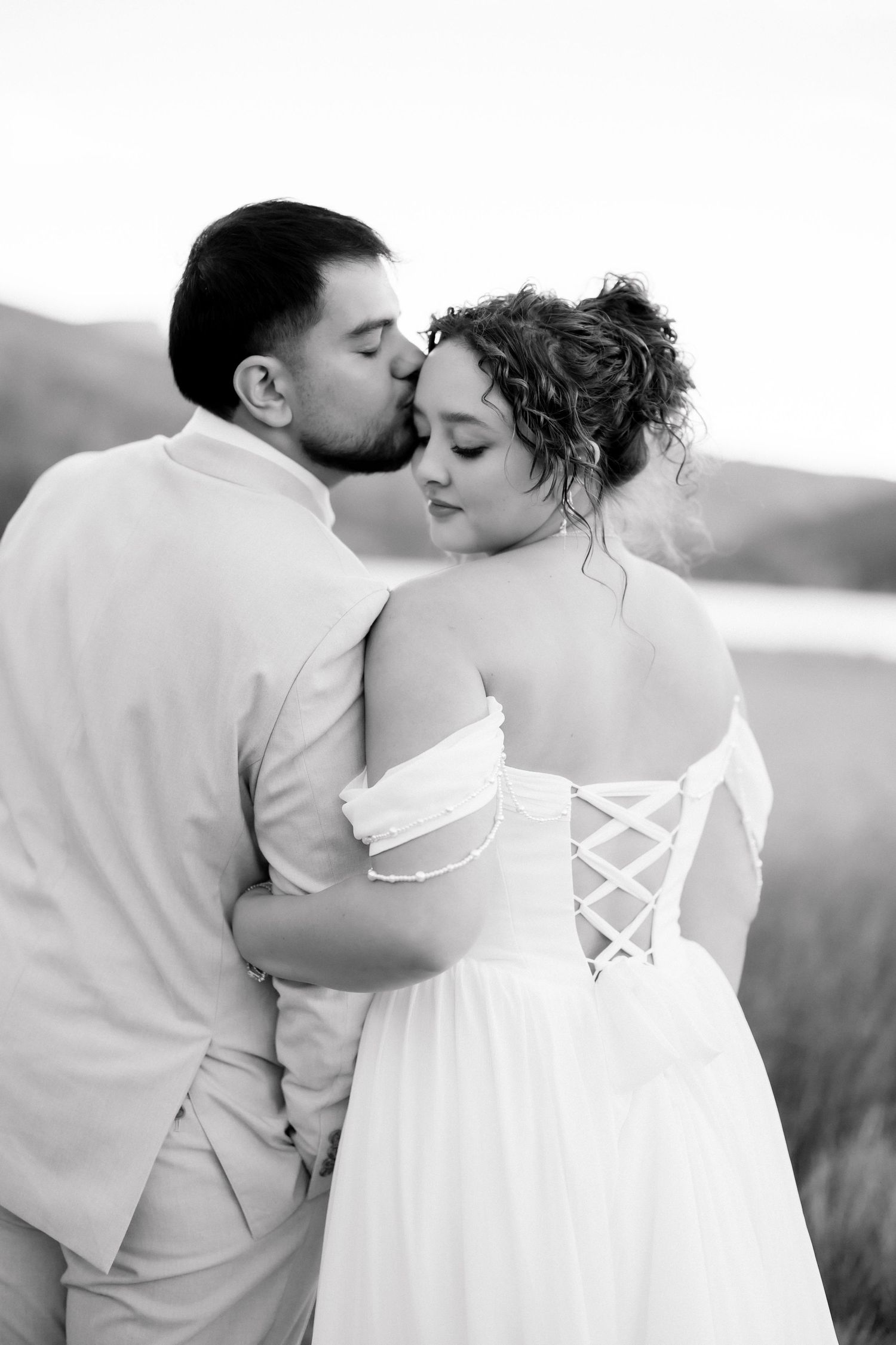 Black and white image of groom kissing bride on cheek, highlighting the corset back detail of her bridal gown.
