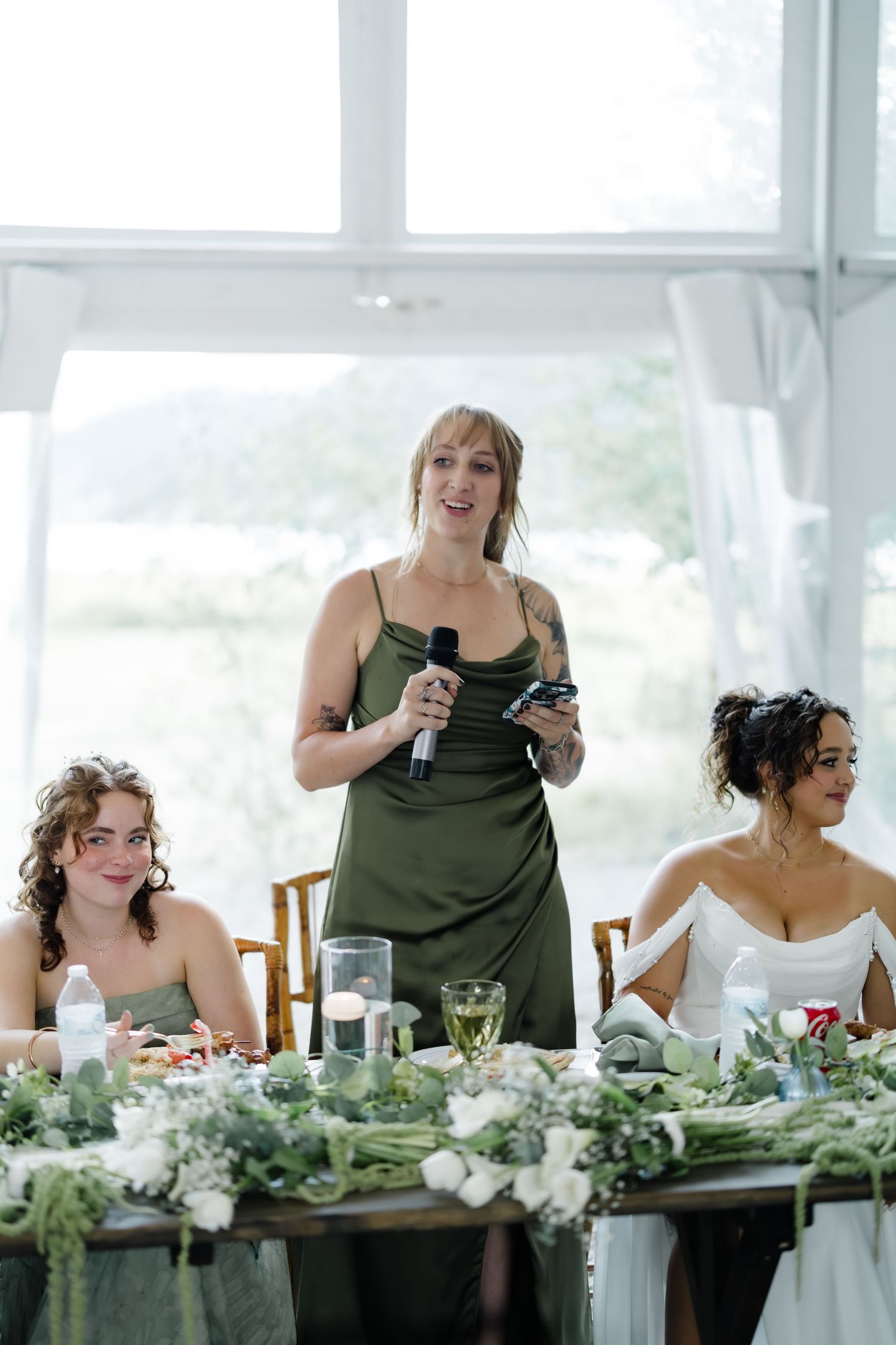 Bridesmaid in olive green dress gives a heartfelt speech at wedding reception head table.