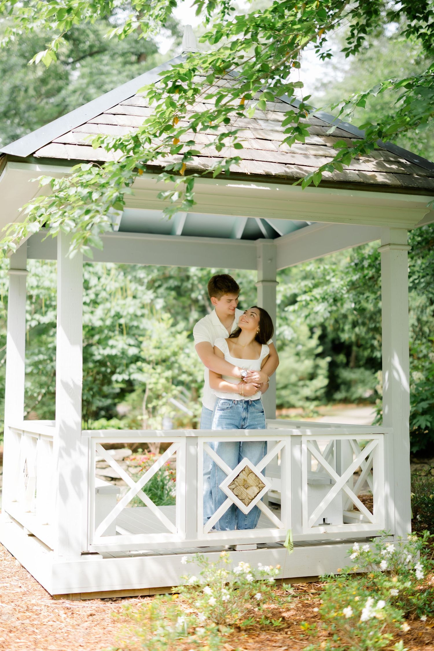 Couple embracing romantically in a white gazebo surrounded by lush green trees in a garden setting.