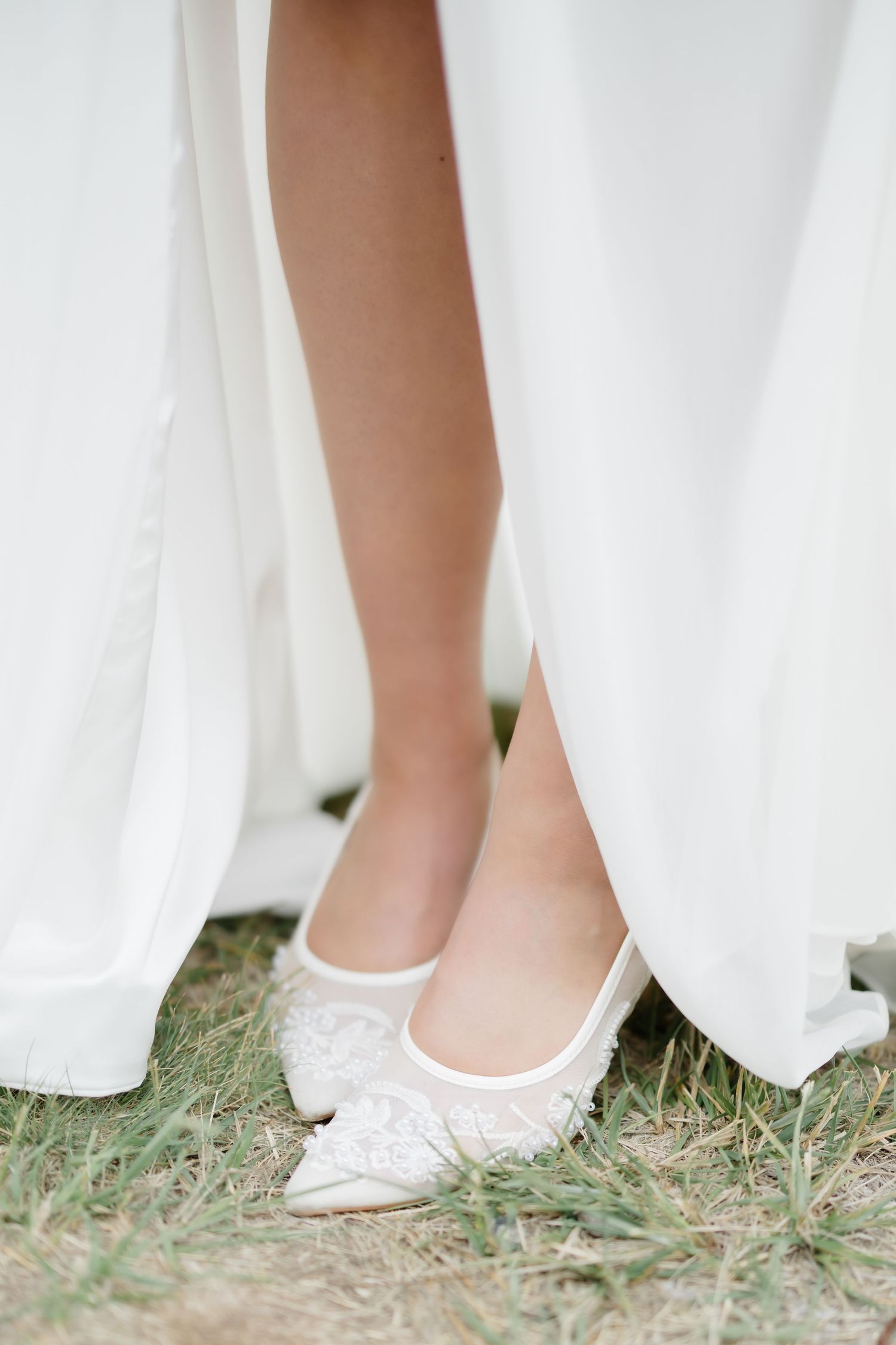 Bride wearing white lace pointed-toe flats on grass, peeking beneath a flowing white wedding gown.