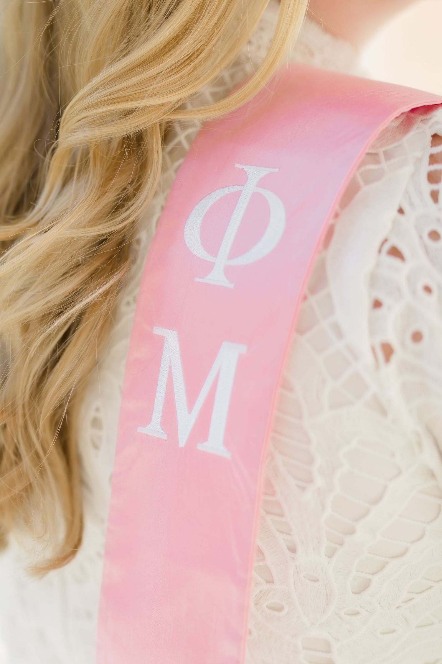 Close-up of pink Phi Mu sorority graduation sash worn over white lace dress by blonde graduate.