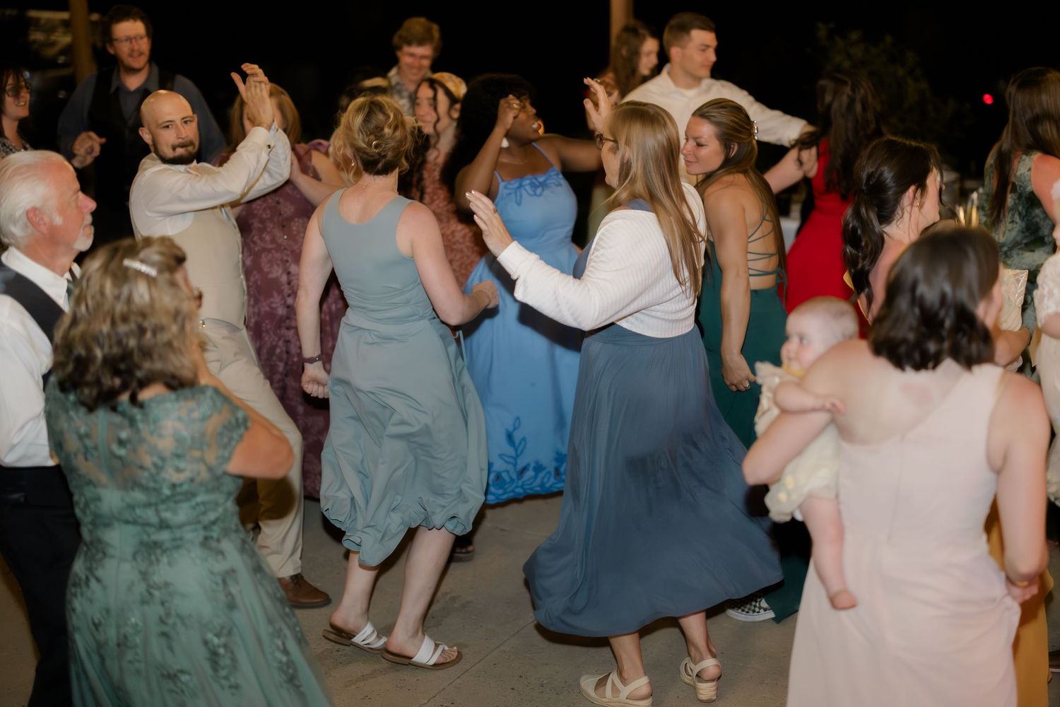 Guests dancing joyfully at a wedding reception, wearing elegant dresses in blue, green, and white tones.