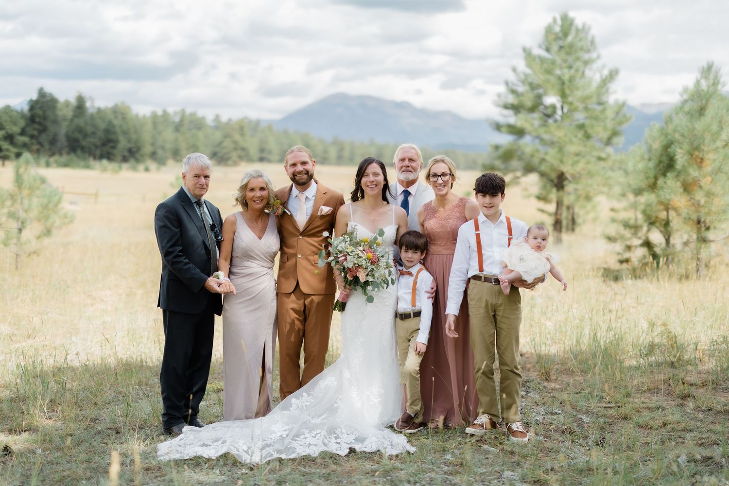Wedding party poses in rustic mountain meadow, bride in white gown, groom in tan suit, surrounded by family.