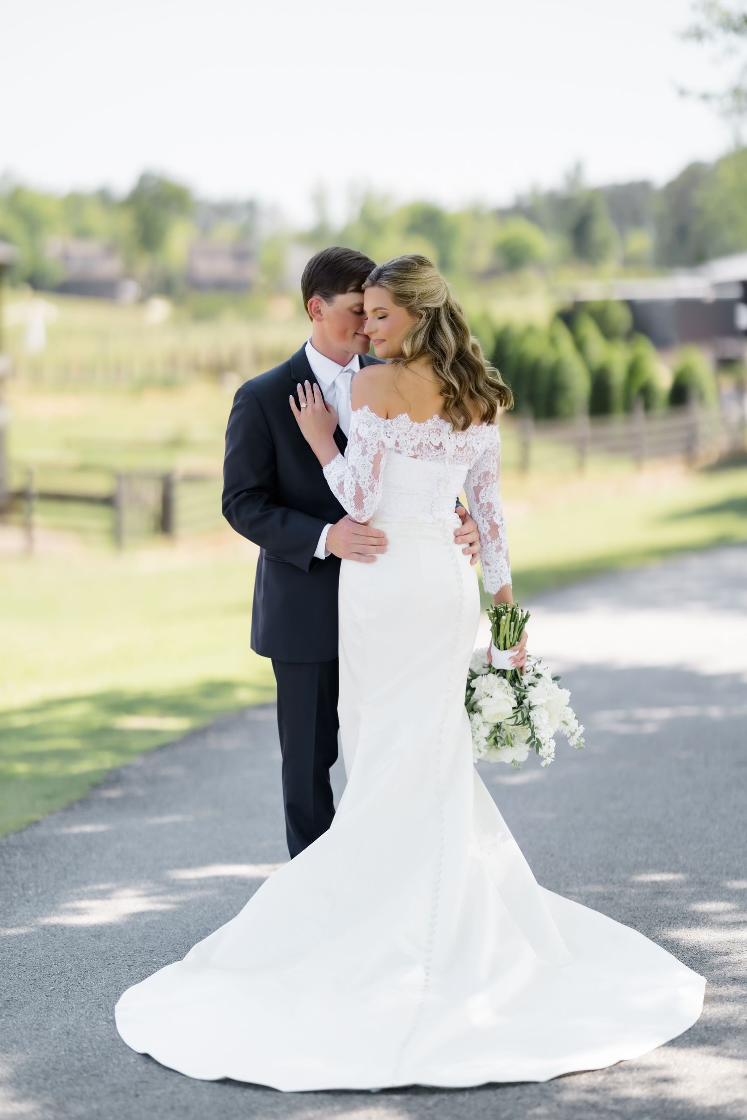 Bride in lace gown and groom in navy suit share a romantic embrace on a path at an outdoor wedding venue.