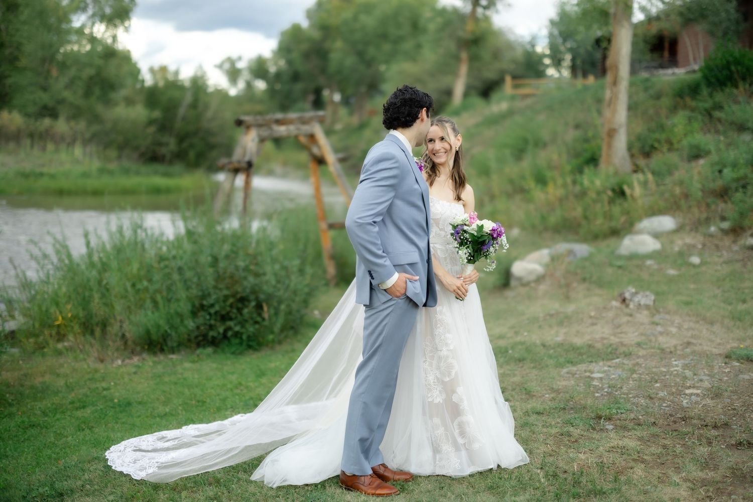 Bride and groom pose by a serene pond, bride in lace gown with long train holding purple bouquet.