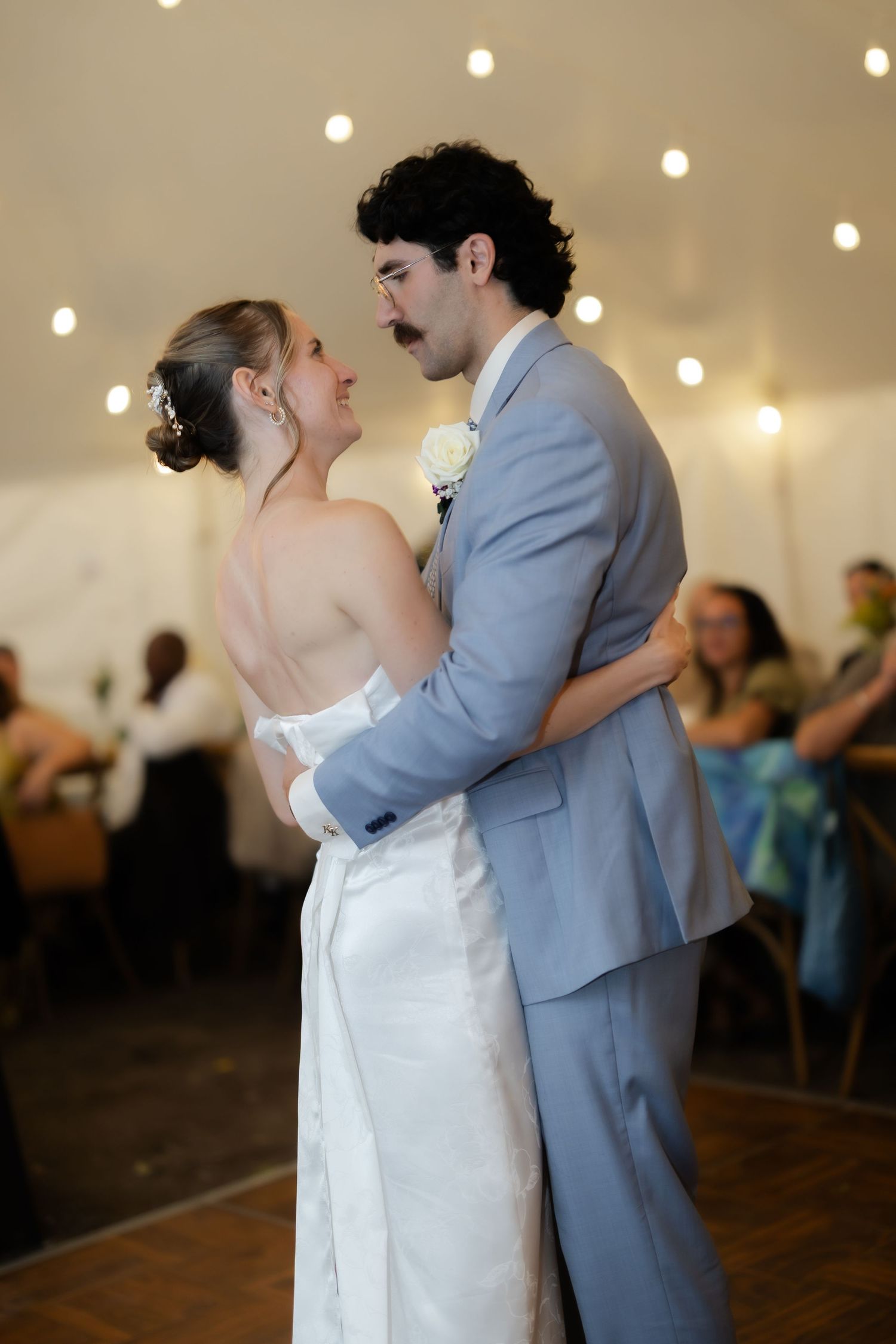 Bride and groom share their first dance under string lights at a wedding reception in a white tent.