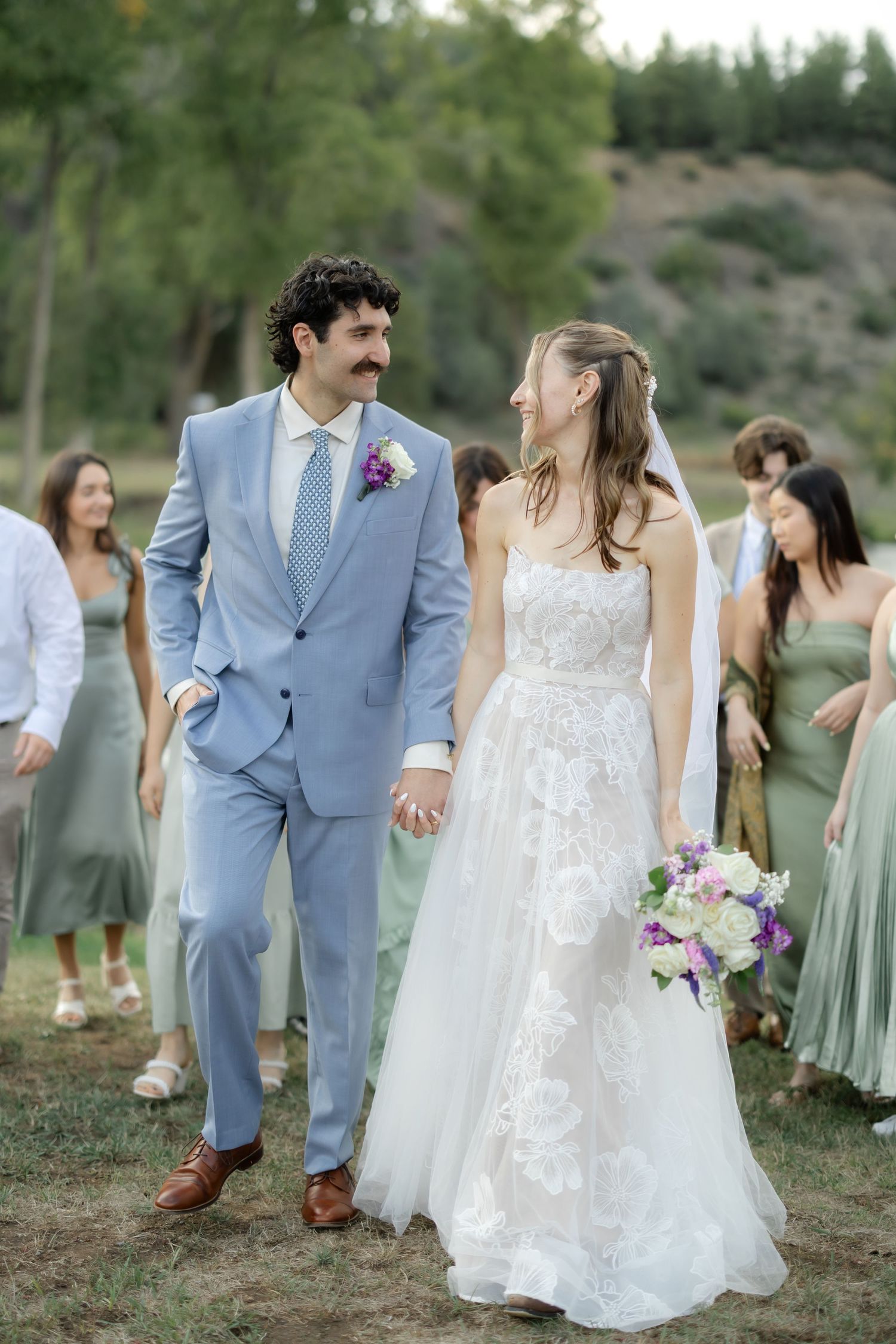 A bride and groom walk hand in hand outdoors, with bridesmaids in sage green dresses following behind them.