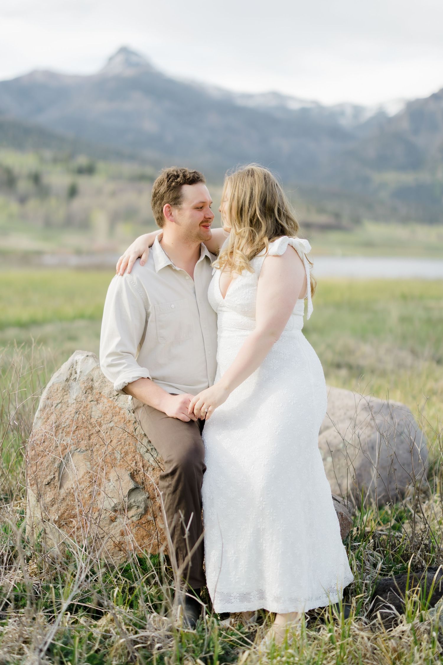 Couple sharing a romantic moment leaning against a boulder in a mountain meadow, woman in white dress.