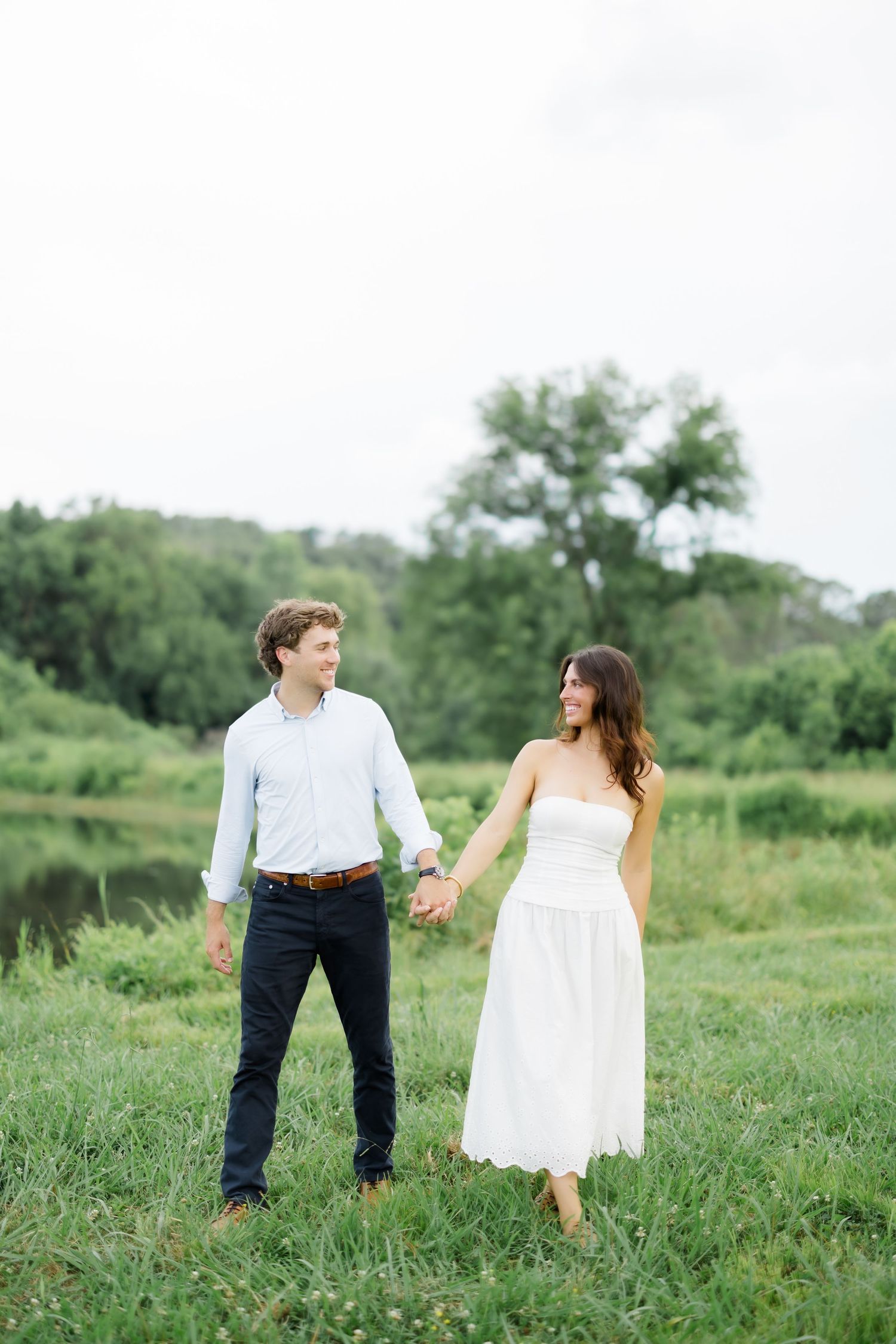 A smiling couple holds hands while walking through a green meadow near a serene pond on a cloudy day.