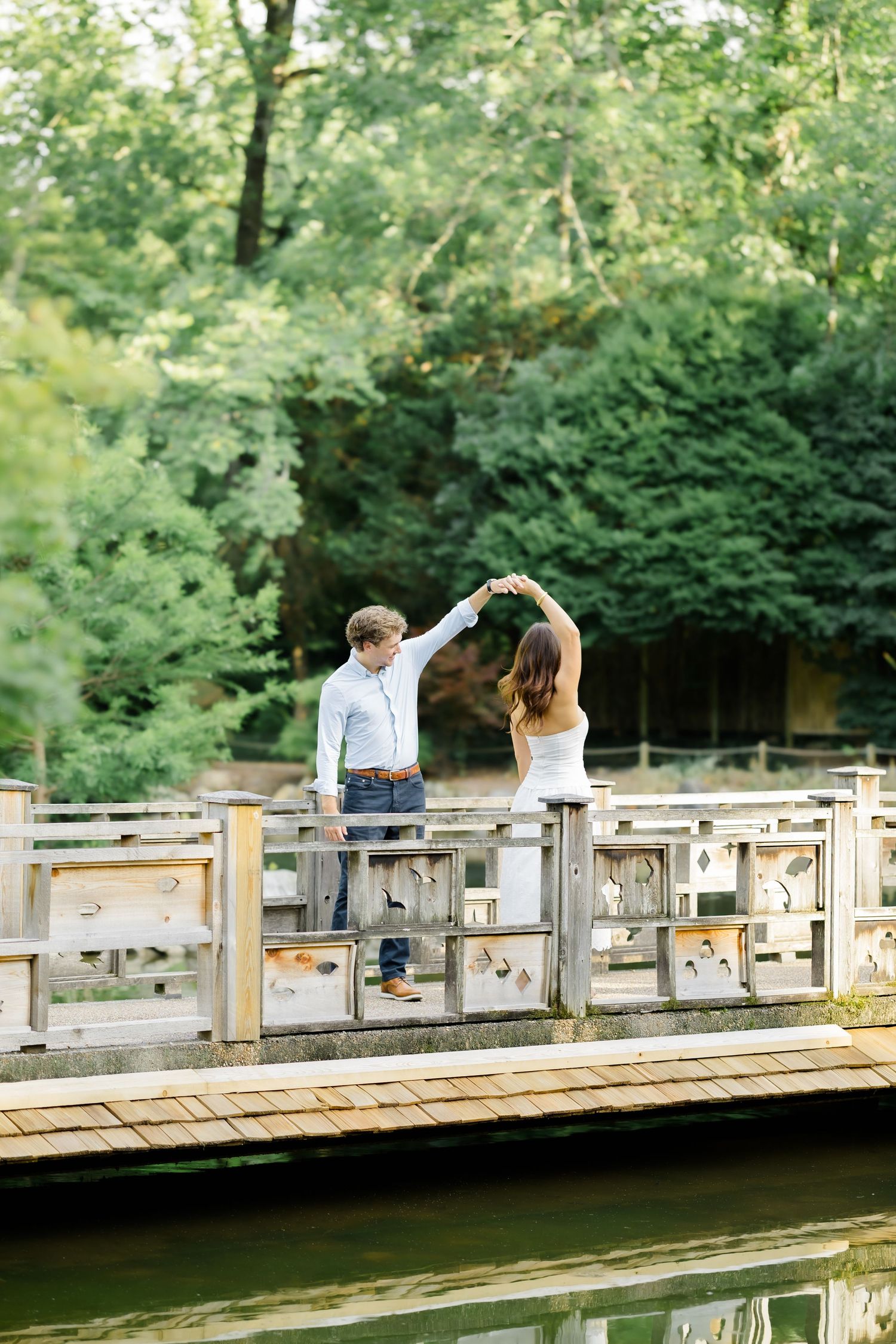 Couple dancing on a wooden bridge over a pond surrounded by lush green trees.