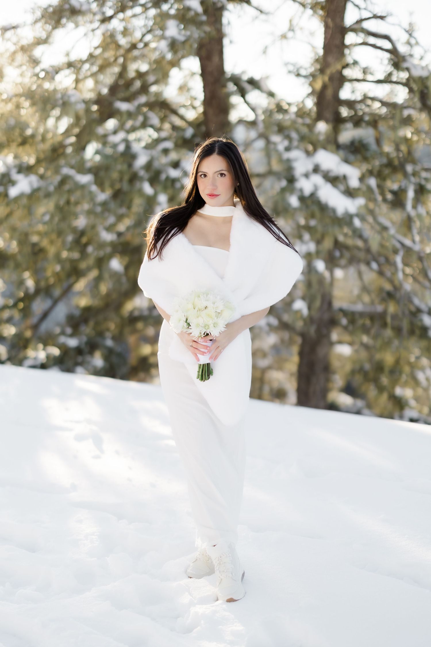 Elegant bride in white dress and fur wrap holds bouquet in a snowy winter forest.