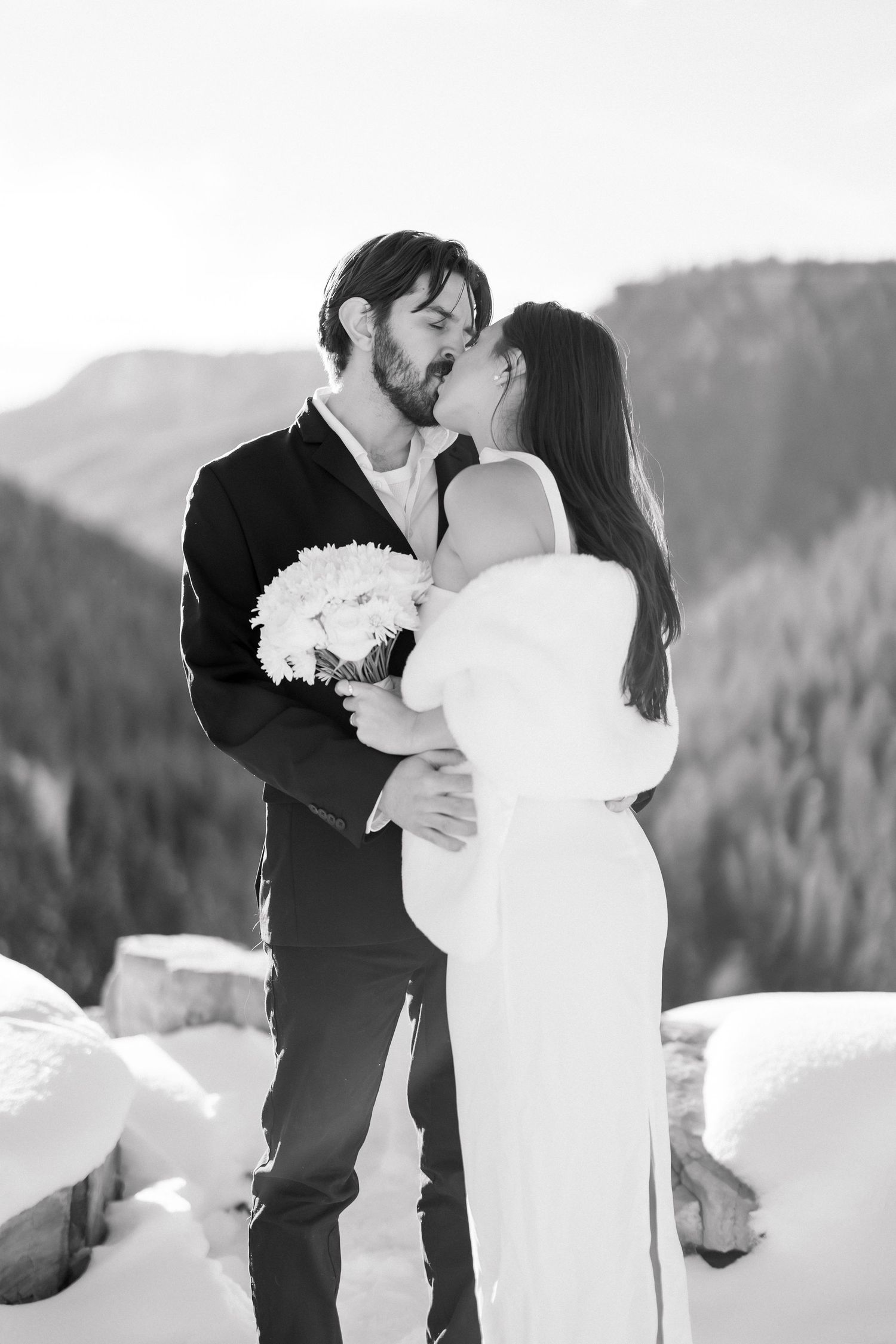 Black and white photo of bride and groom sharing a passionate kiss in snowy mountain setting.