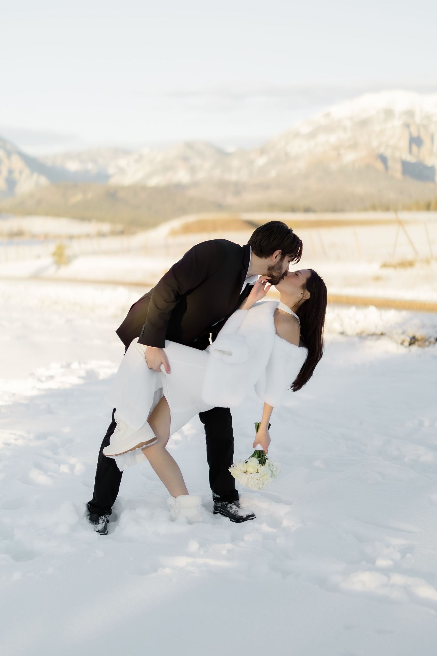 Groom dips bride for a kiss in snowy mountain landscape during romantic winter wedding photo.
