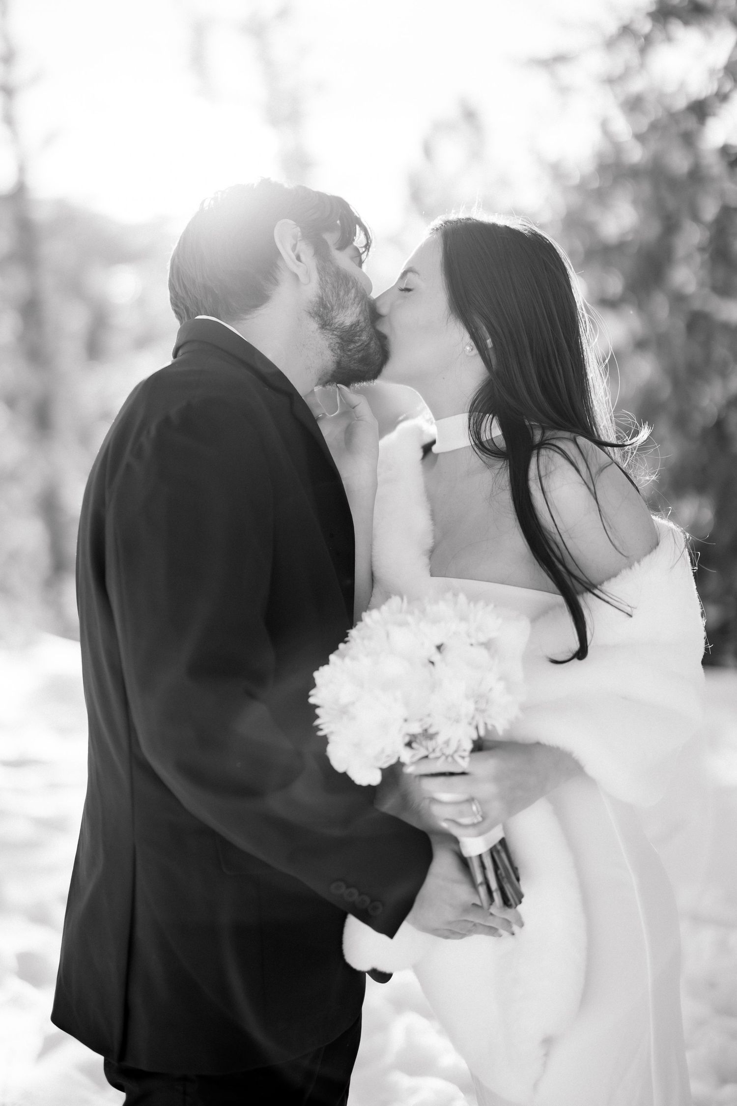 Black and white photo of groom kissing bride holding white bouquet in sunlit snowy forest.