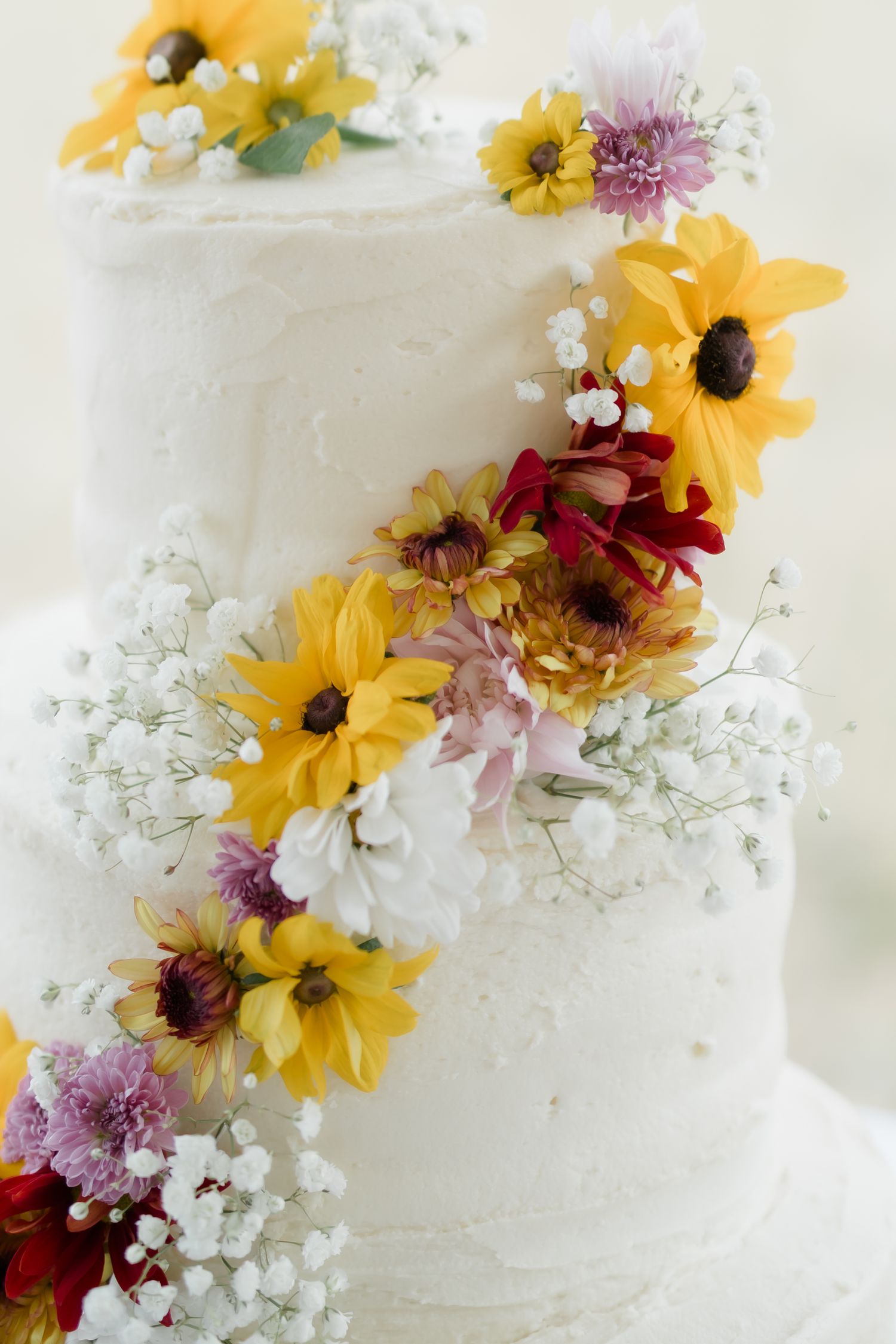 White two-tier wedding cake decorated with colorful sunflowers, daisies, and baby's breath cascading down the layers.